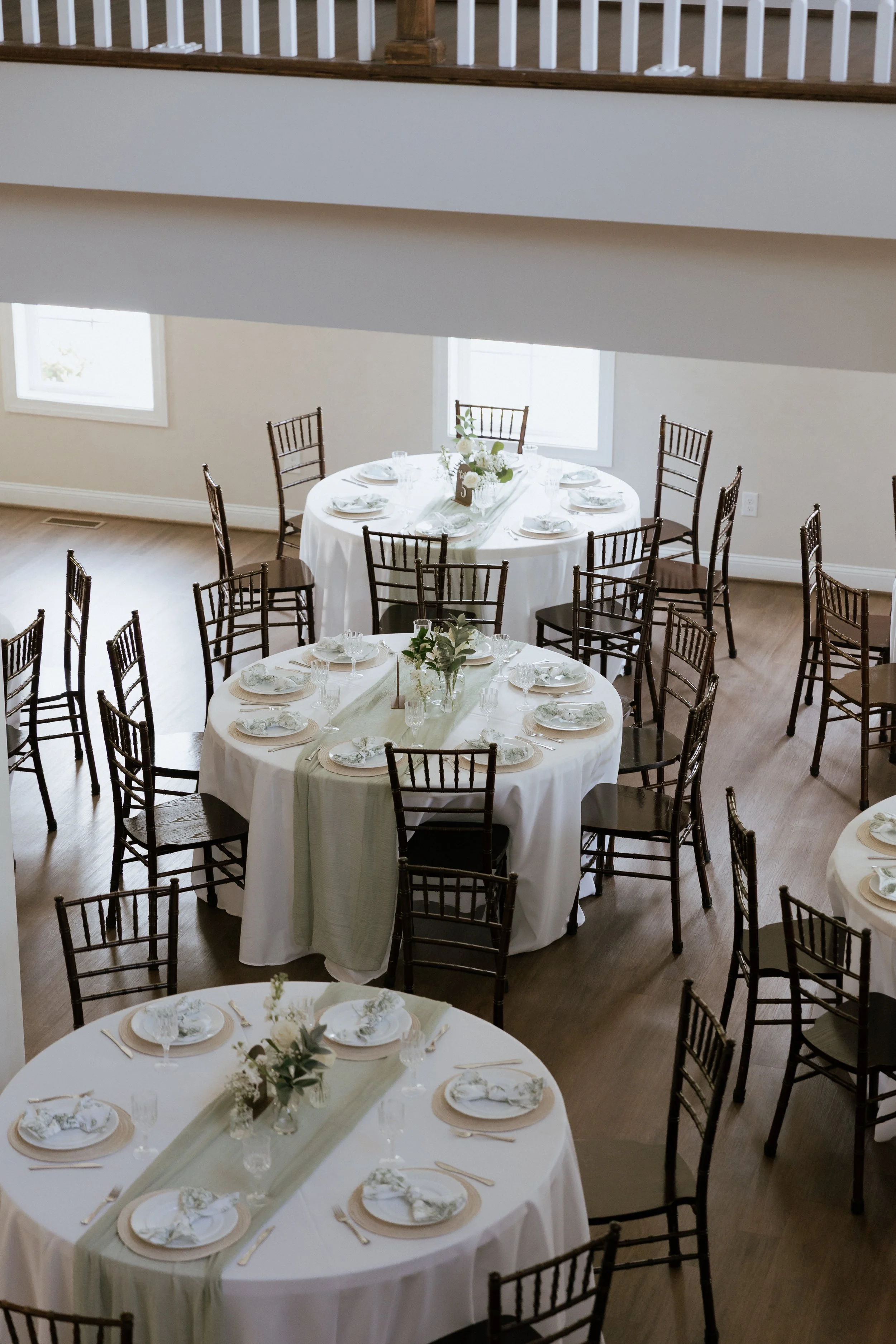 An elevated photo of a wedding reception's table settings and details, where round tables are decorated with white and green florals, natural wood tones, and sage green cheese cloth runners