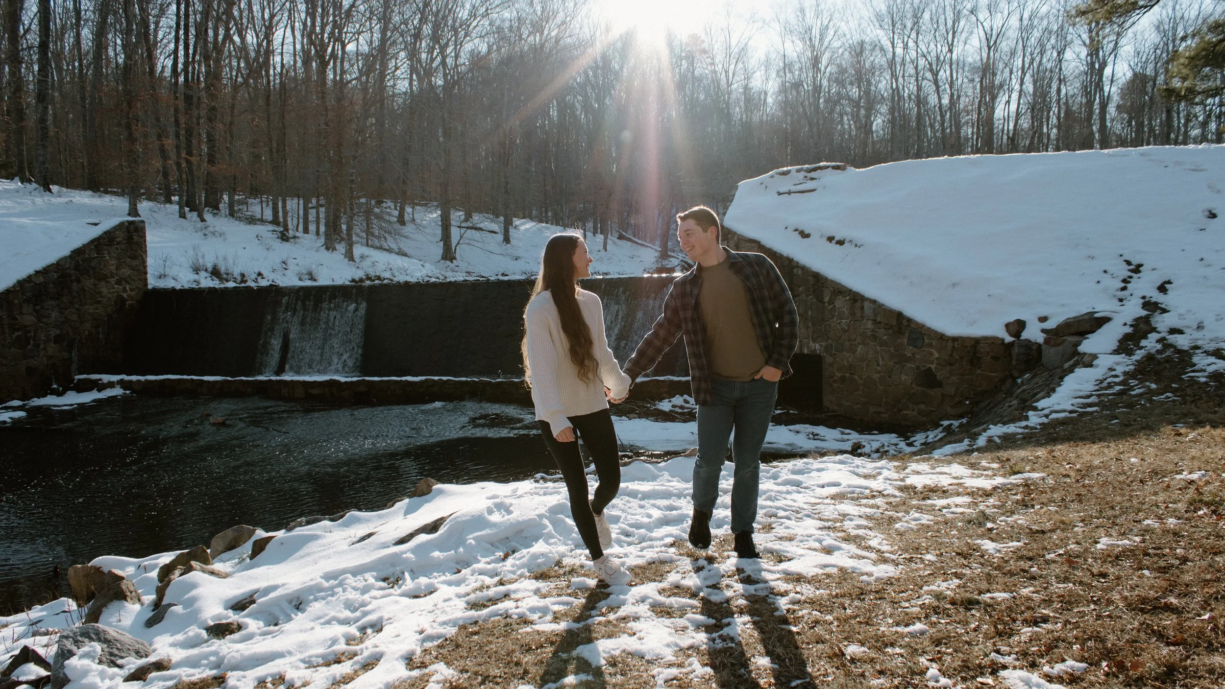 A couple holding hands and walking in the snow near a small waterfall with snow-covered banks on a sunny day.