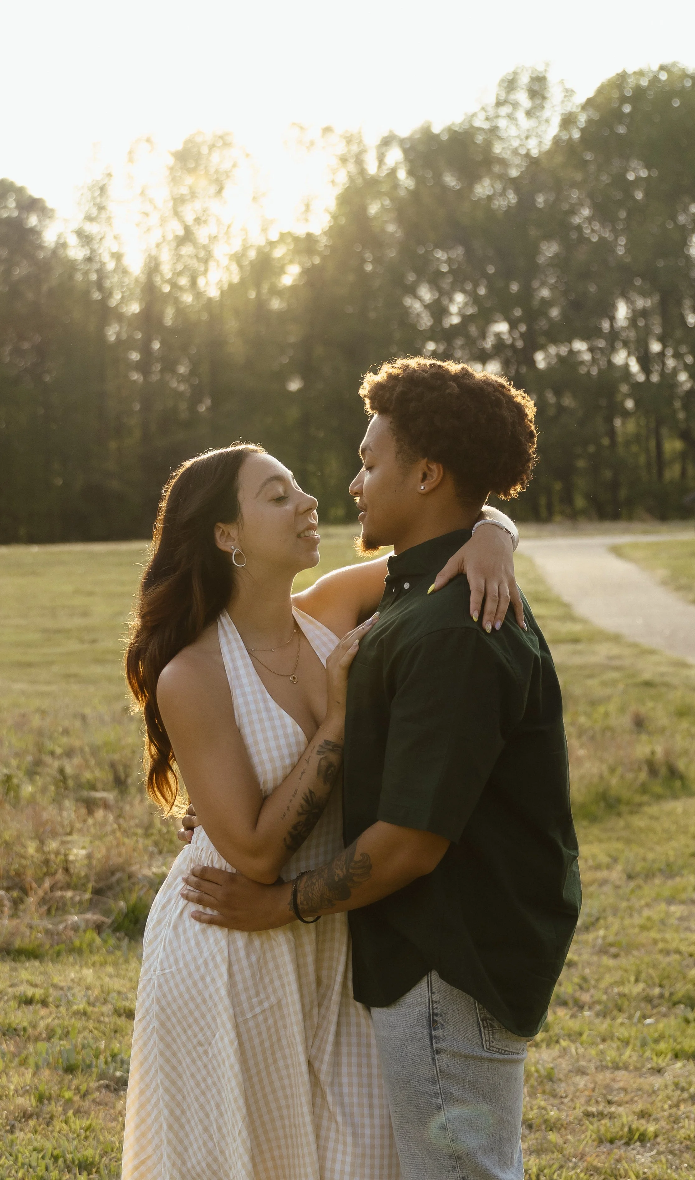 A couple embracing outdoors during sunset, facing each other affectionately in a field with trees in the background.