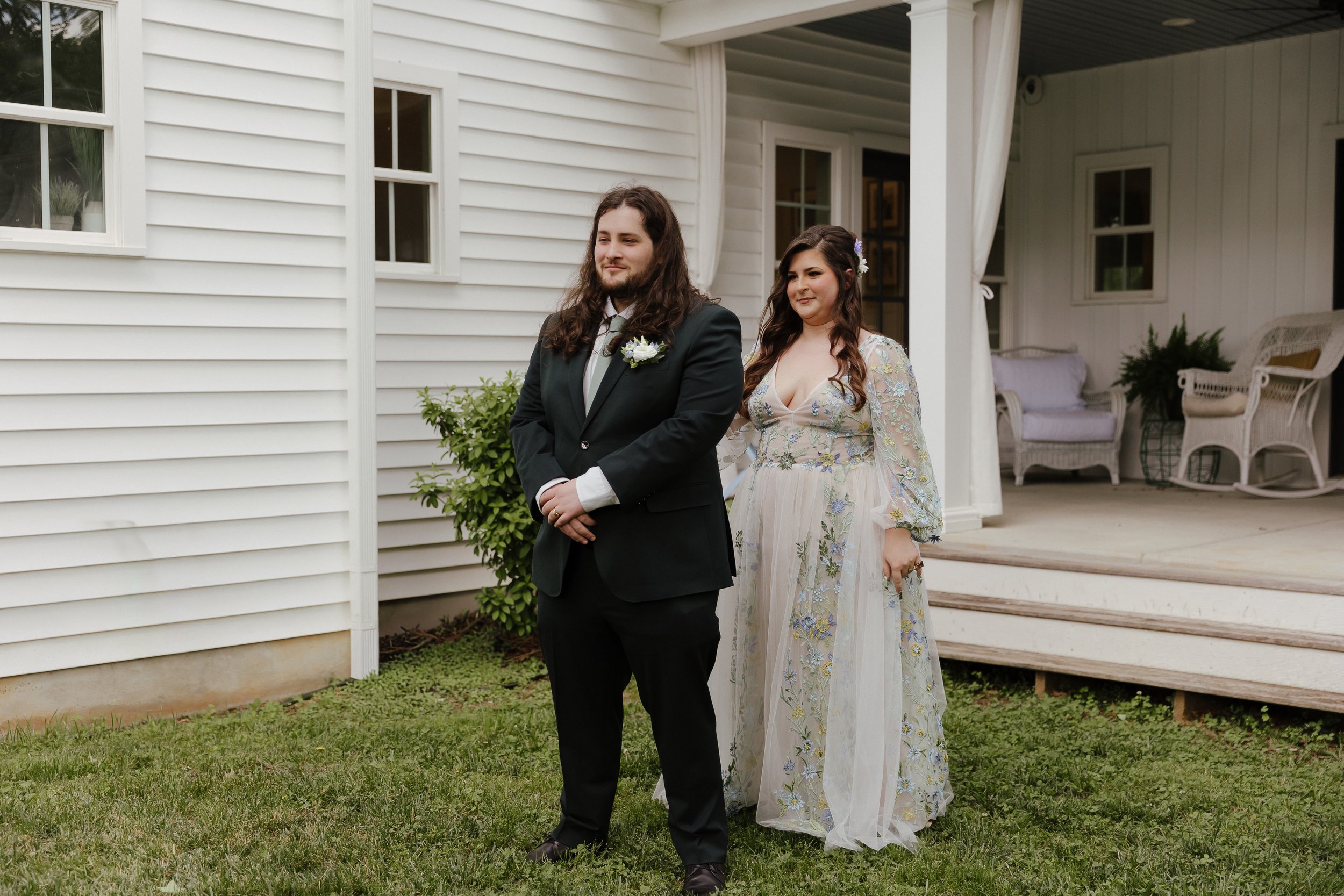 A photo of a couple, where the woman is standing behind the man in a wedding gown that has colorful, intricate lace details. The man is in a dark green suit, with a sage green tie.