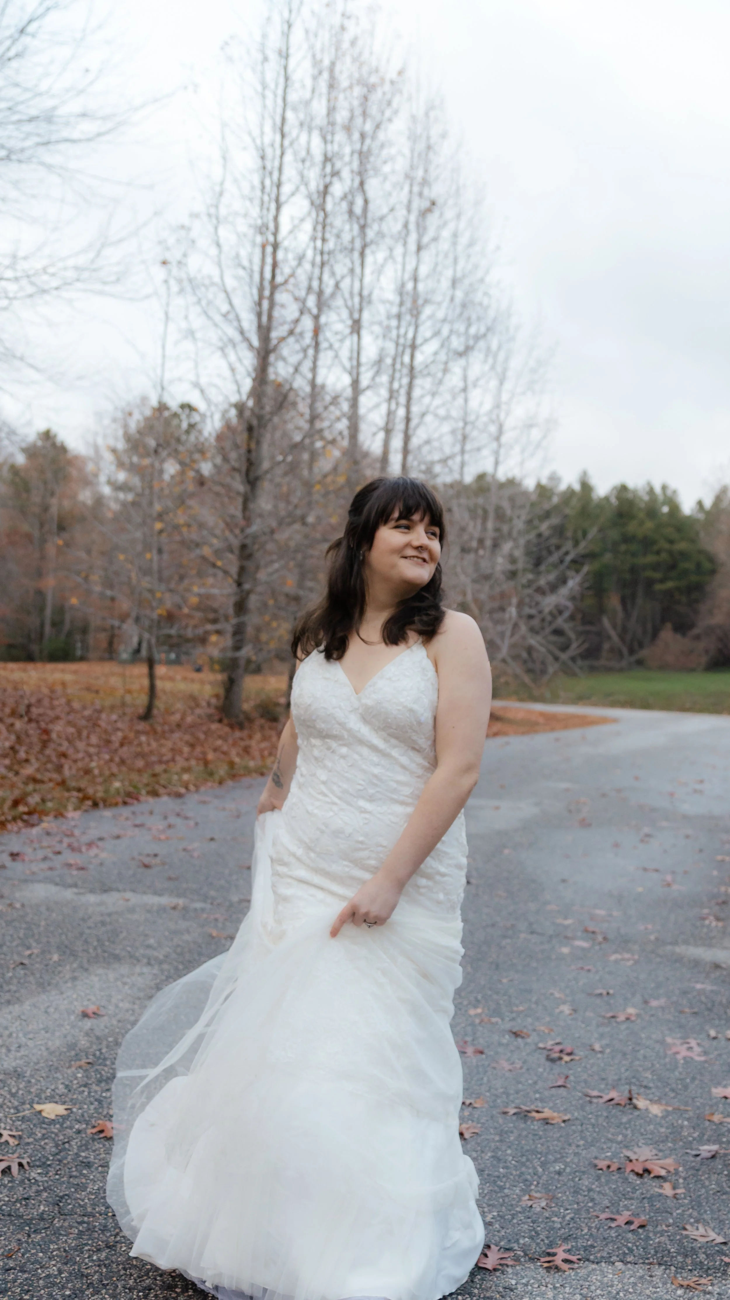 A woman in a wedding dress standing outdoors on a paved path surrounded by leafless trees and fallen autumn leaves.