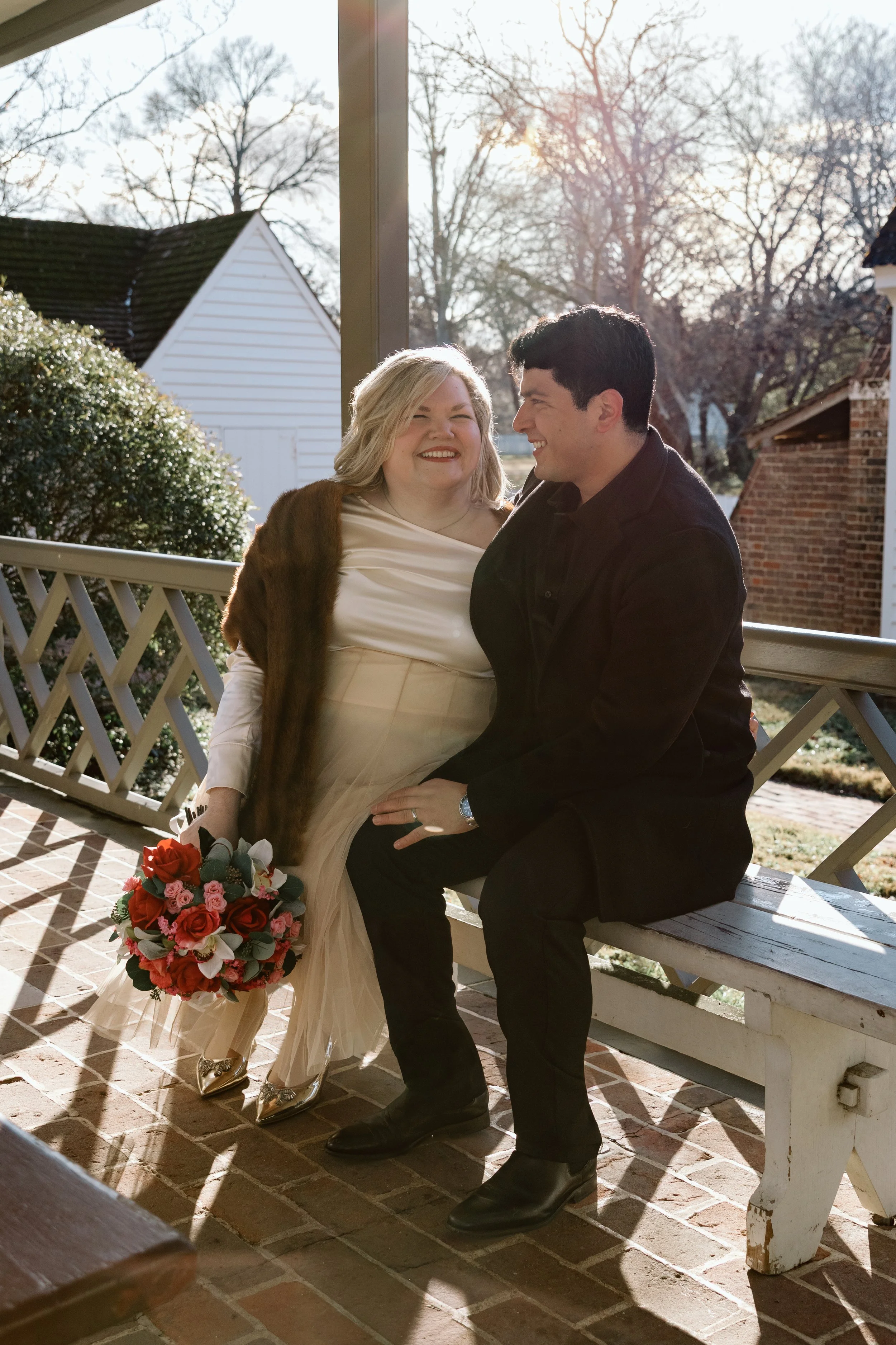 A couple sitting on a bench outside, smiling and looking at each other, with the woman holding a bouquet of pink, red, and white roses, during a sunny day.
