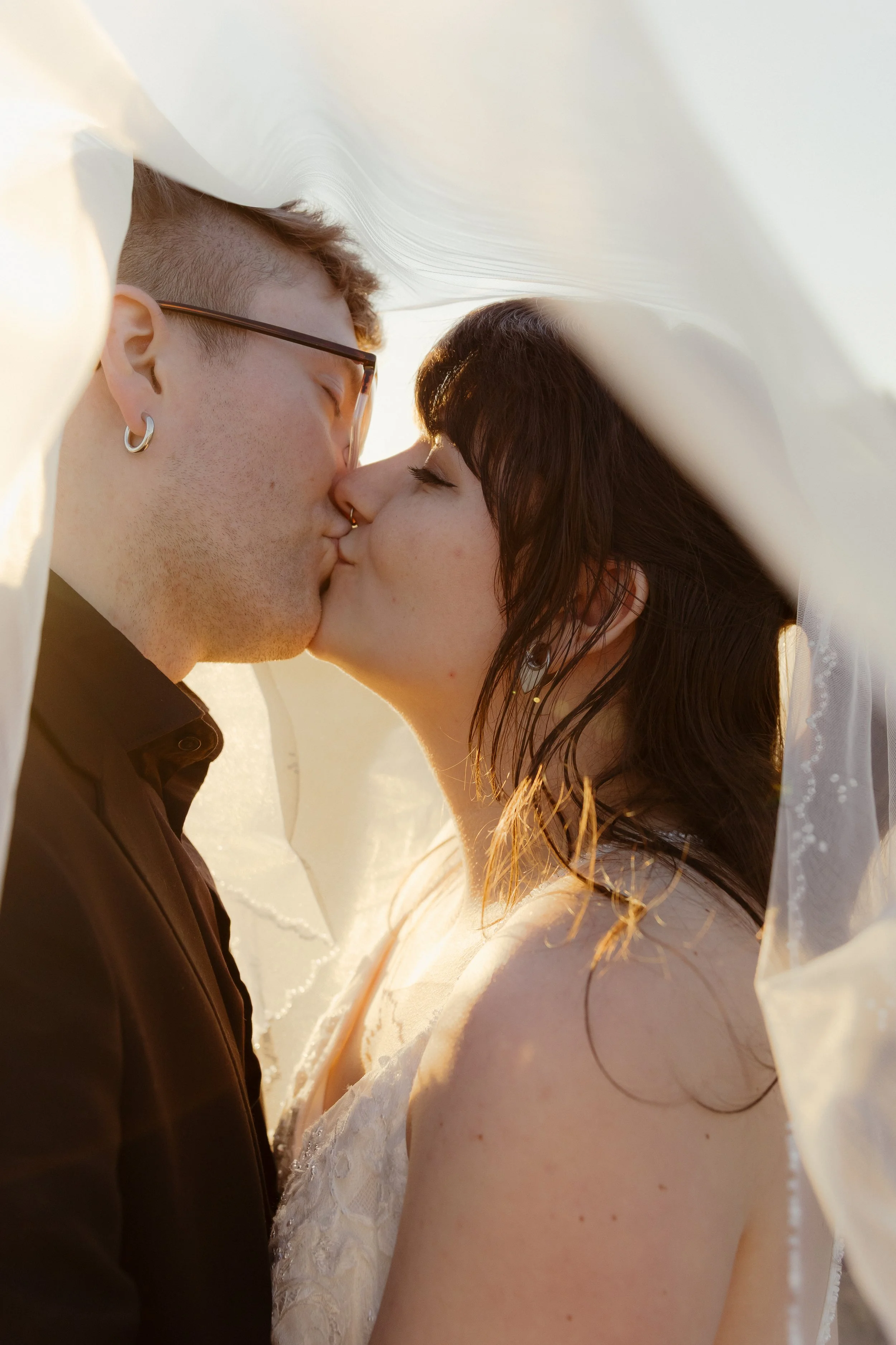 A couple sharing a kiss under a sheer white fabric, with the sunlight illuminating their faces. The man wears glasses and earrings, and the woman has dark hair and earrings.