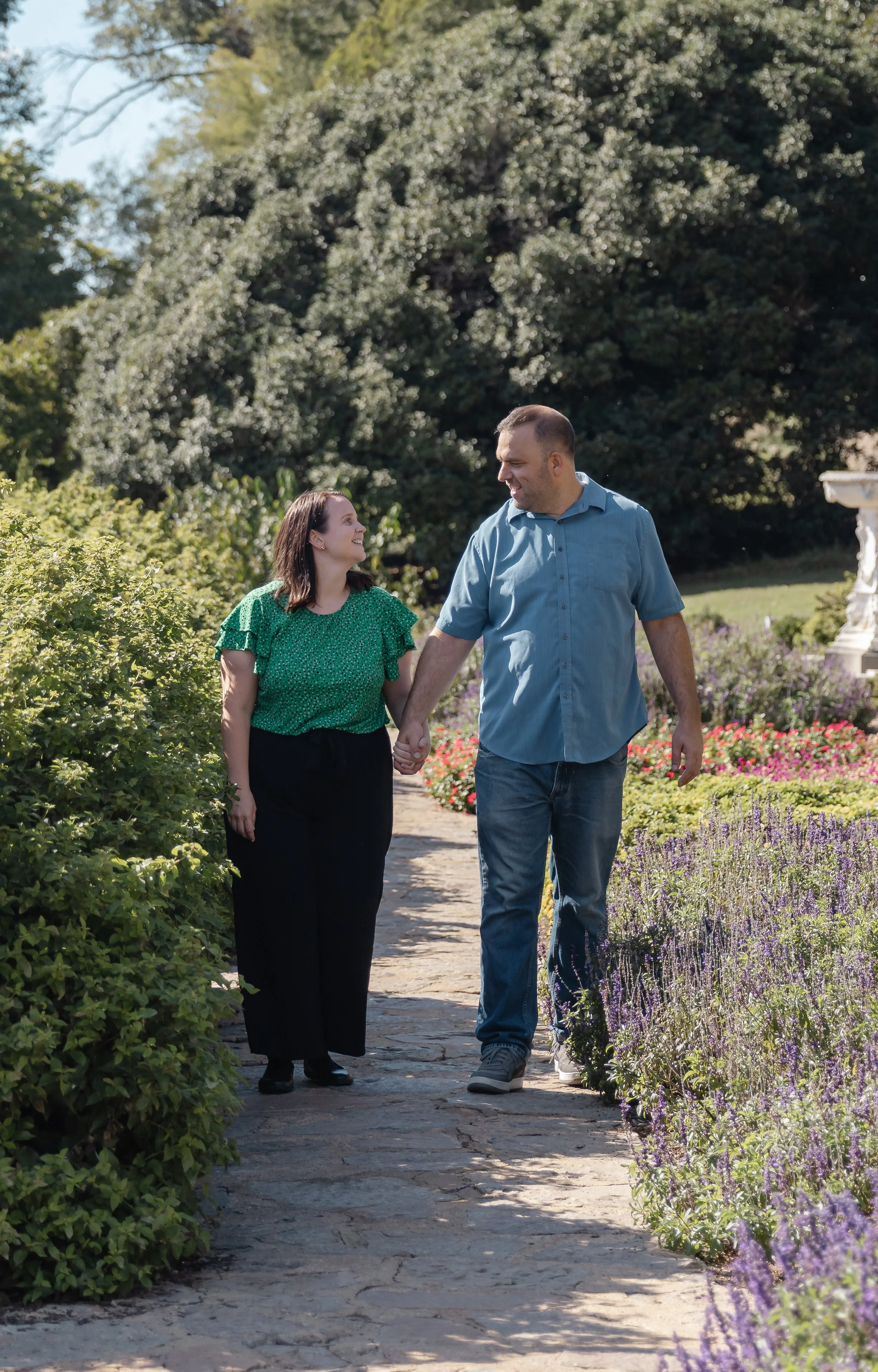 A couple walking hand in hand along a garden path, smiling and looking at each other, surrounded by blooming flowers and lush greenery.