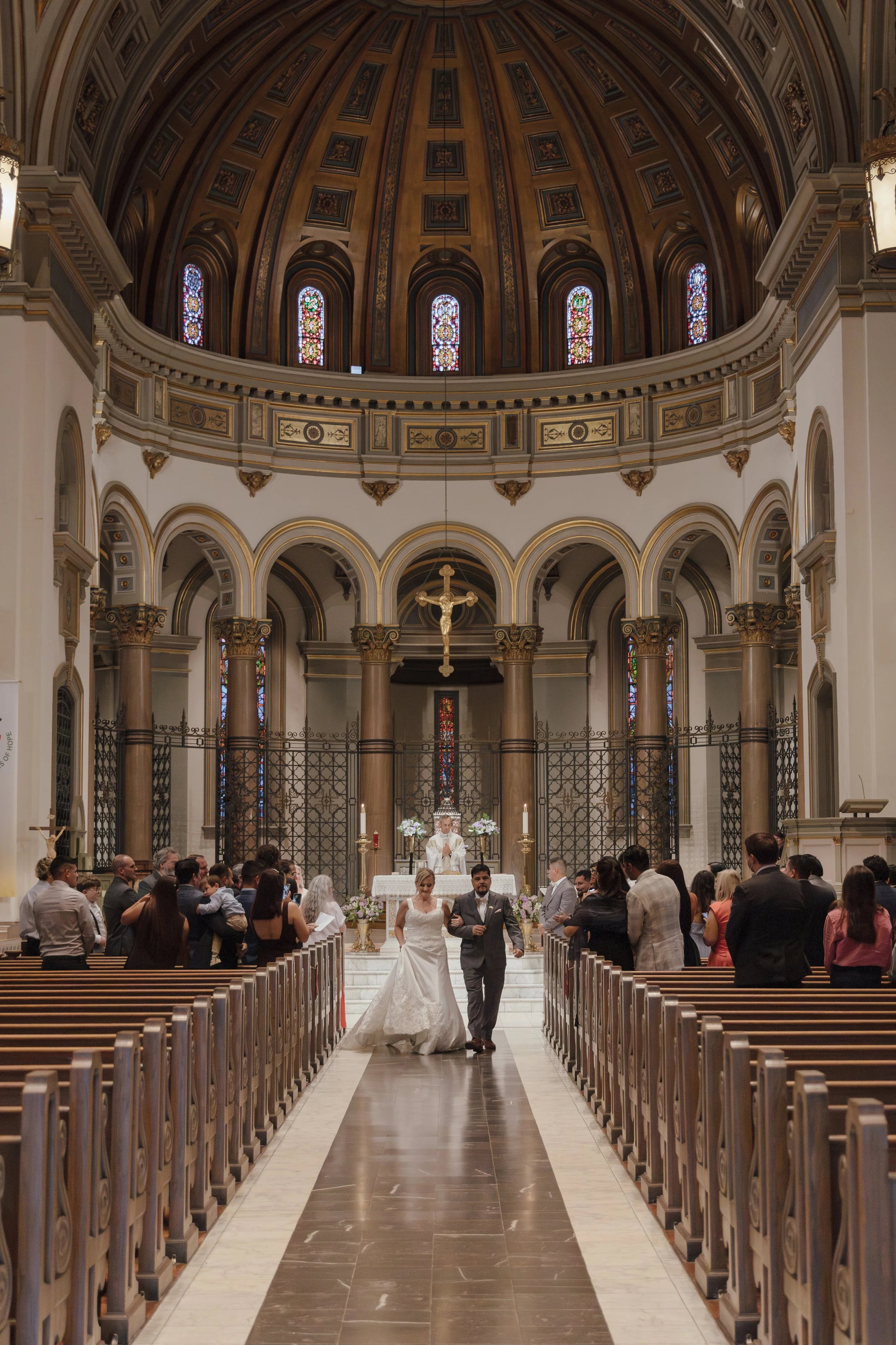 A wedding ceremony taking place inside a grand church with arches, stained glass windows, a dome ceiling, and a crucifix hanging above the altar. The bride and groom are walking down the aisle, surrounded by seated guests.