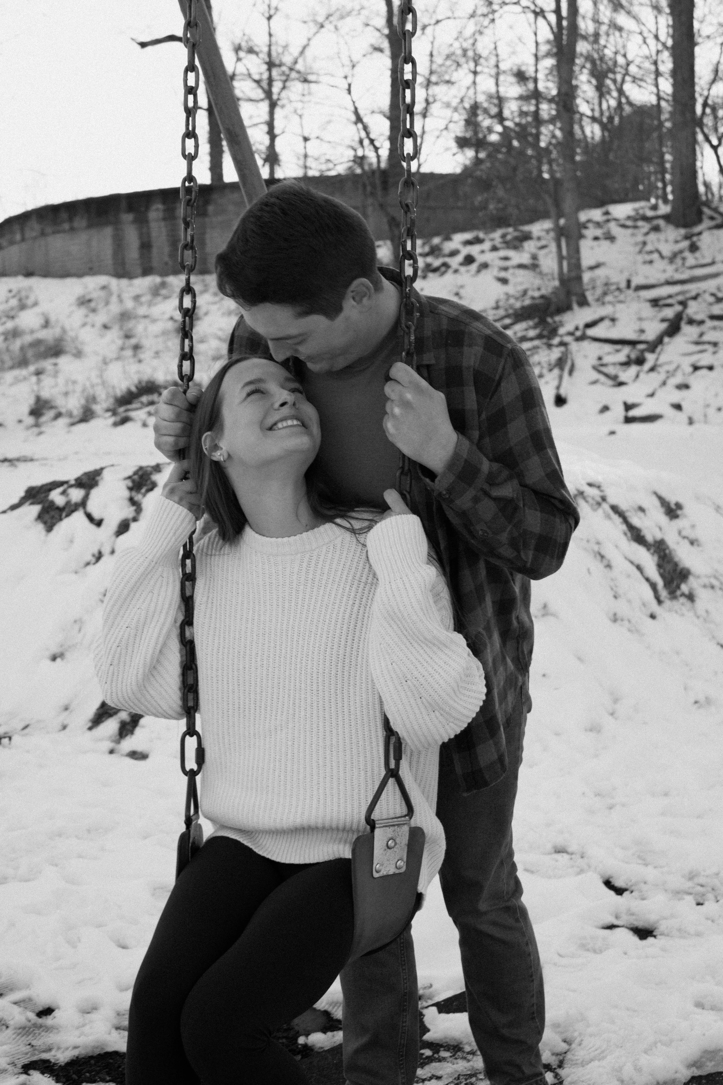 A couple on a swing in a snowy park, smiling and looking at each other.