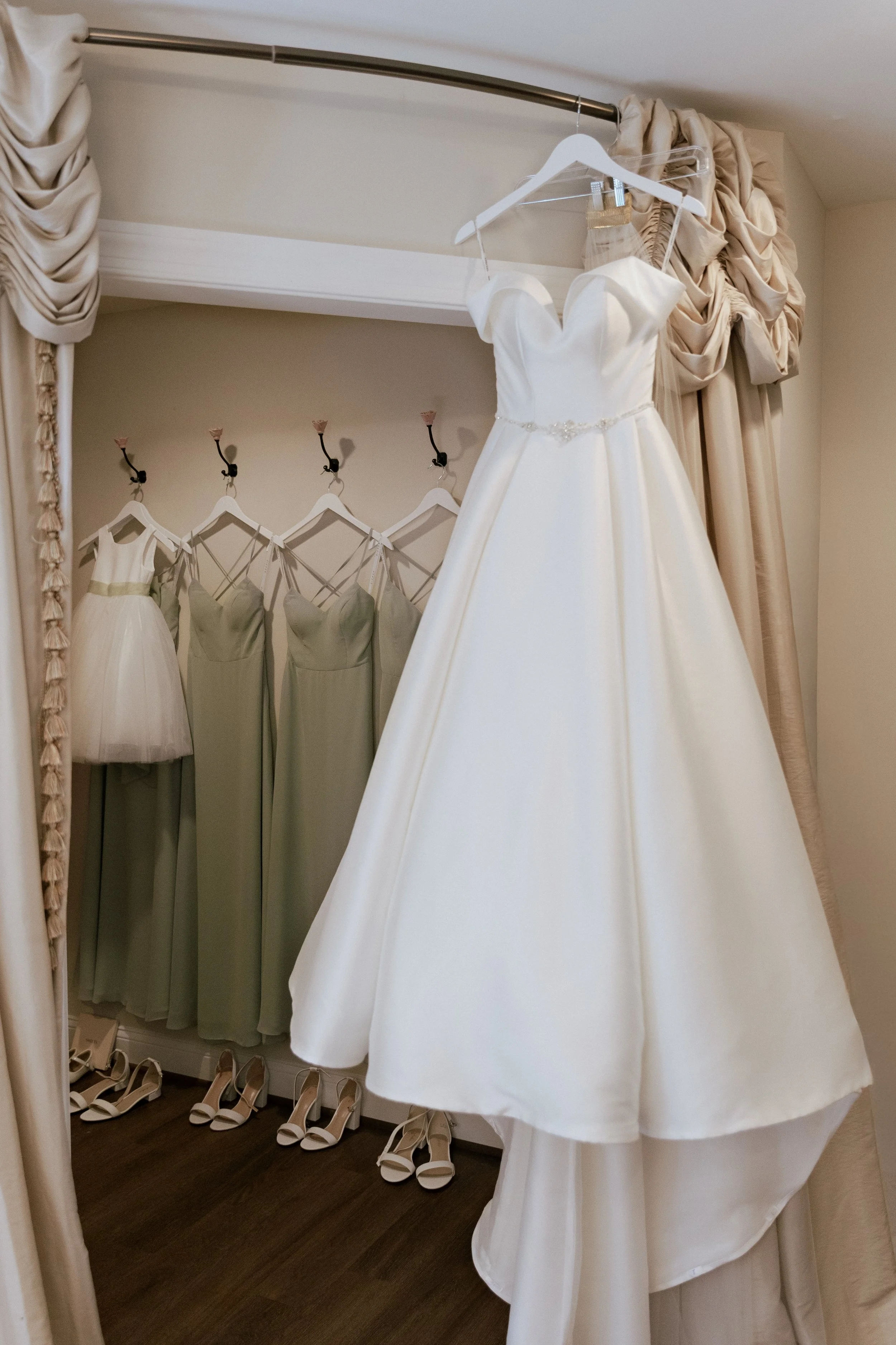White wedding dress hanging on a hanger, with matching shoes on the floor, and more dresses visible in the background.