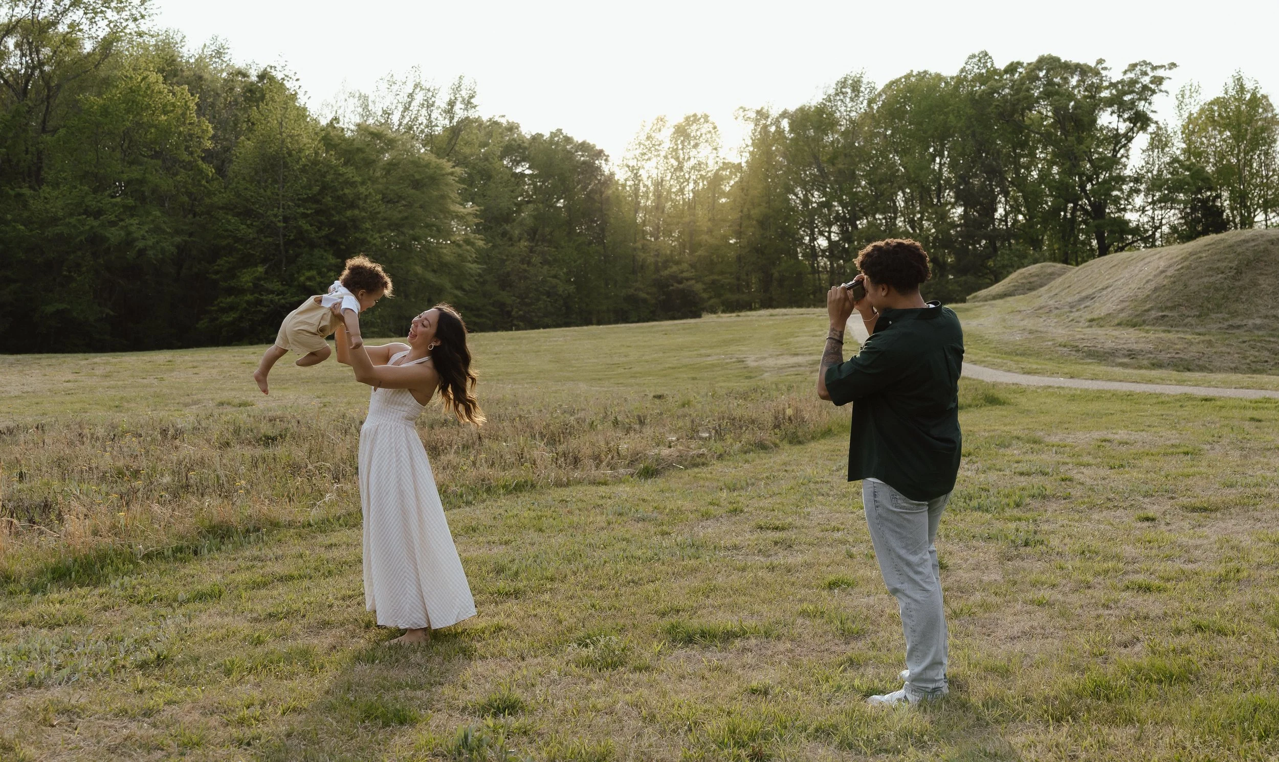 A woman in a white dress holding a young child in a tan outfit in a grassy field with trees in the background, while a man takes pictures of them with a camera.