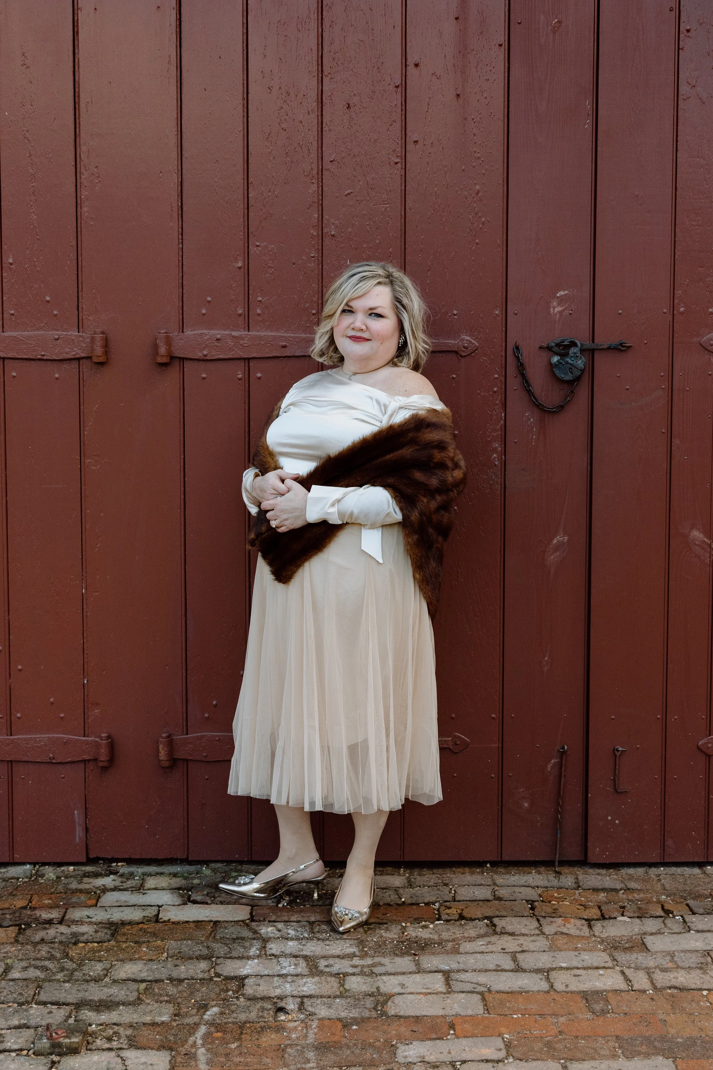 A woman with blonde hair wearing a cream-colored dress, fur stole, and shiny heels standing on a brick ground in front of a large wooden red door with black hardware.