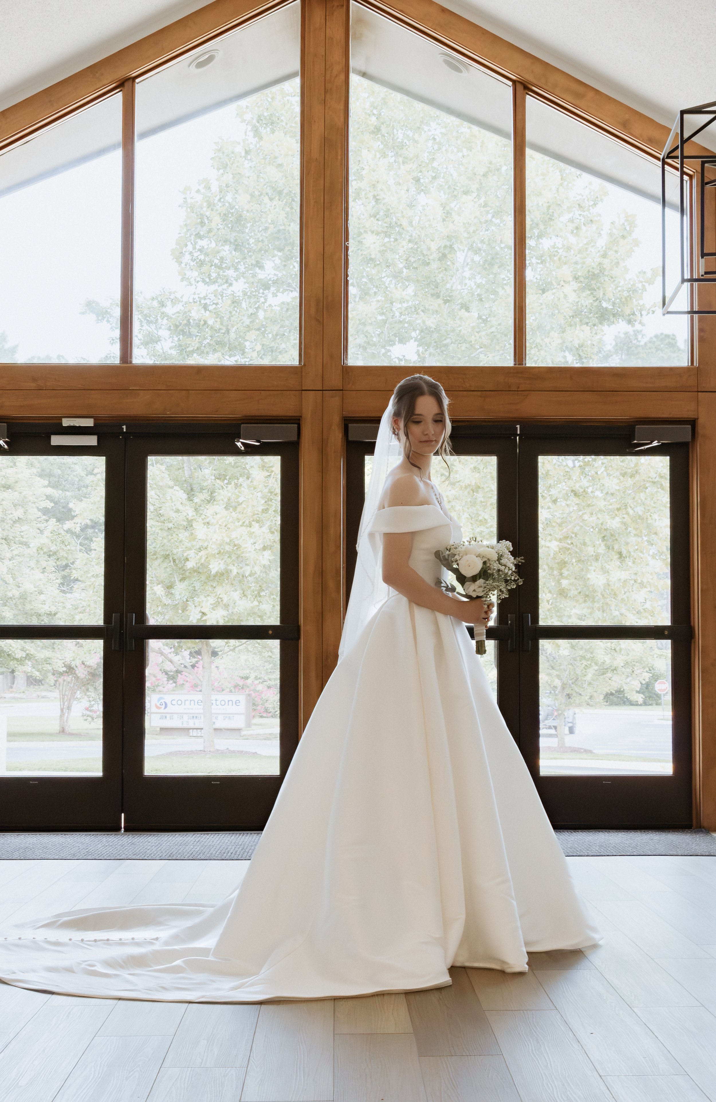 A photo of a bride who is standing in front of large windows, holding a bouquet of flowers in a white ballgown dress and a veil.