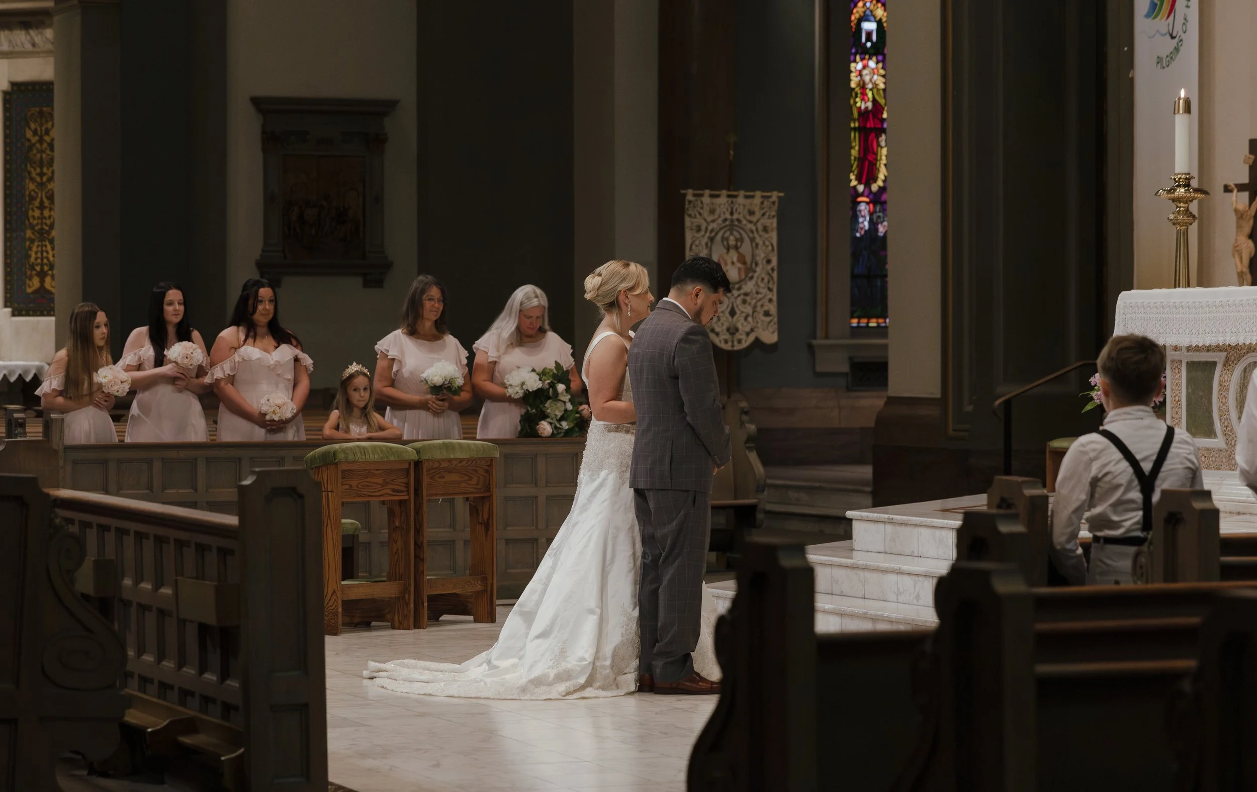 A bride, groom, and wedding party during a church wedding ceremony. The bride in a white gown and the groom in a plaid suit are kneeling at the altar. Bridesmaids hold bouquets in the background.