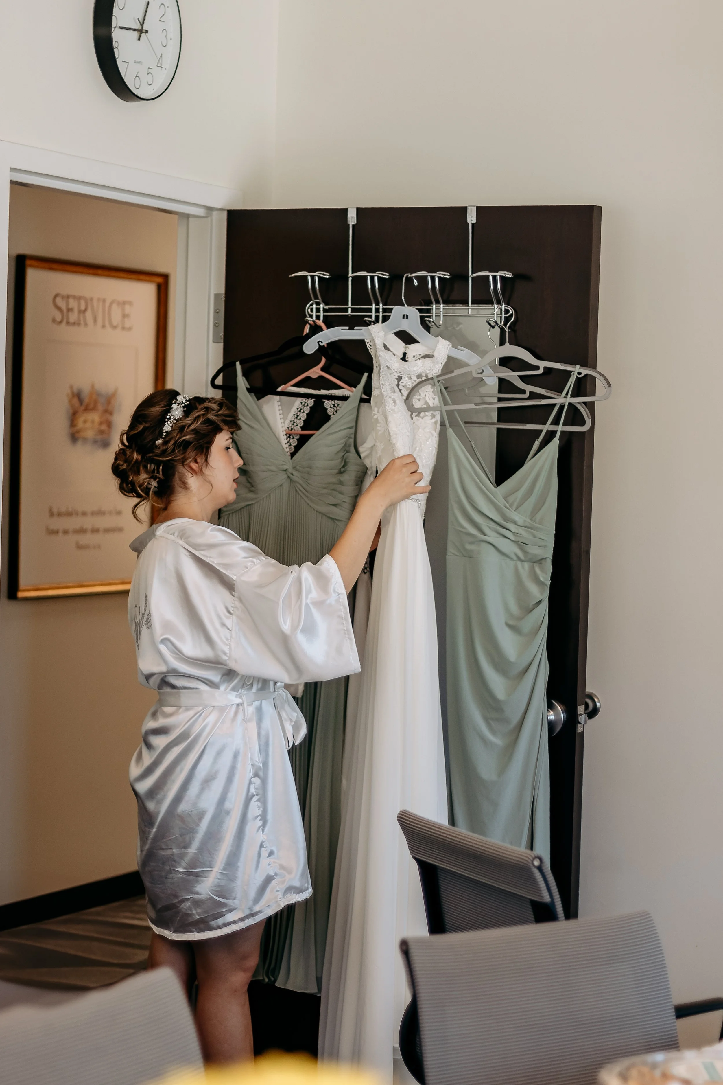 A woman in a satin robe with hair styled in curls and a hair accessory is looking at a white wedding gown hanging on a clothing rack, with bridesmaid dresses in pale green colors also on the rack, inside a room with a wall clock and a framed picture.