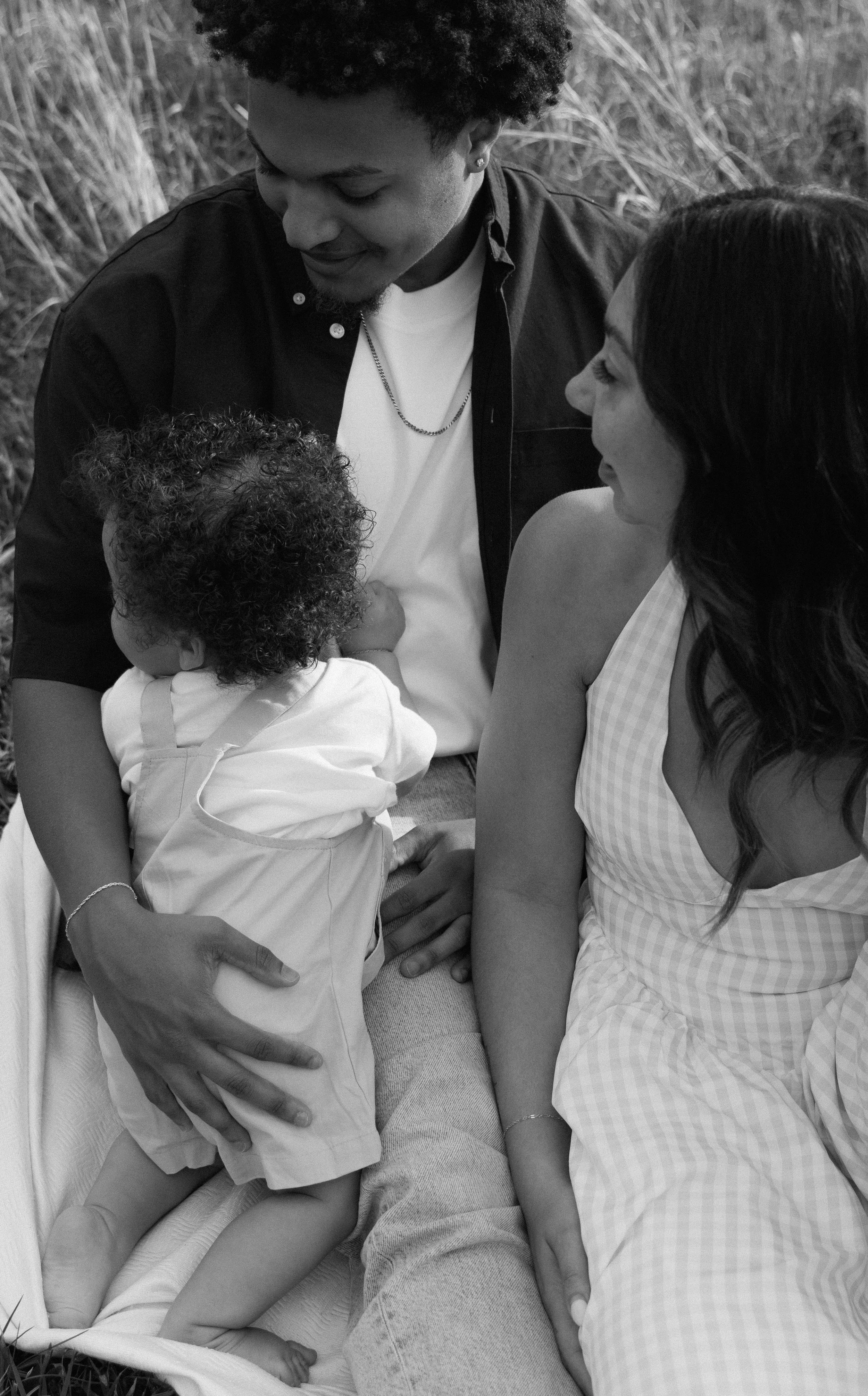A Black woman and a man hold a young child with curly hair outdoors, all smiling with the woman and man looking at the child.