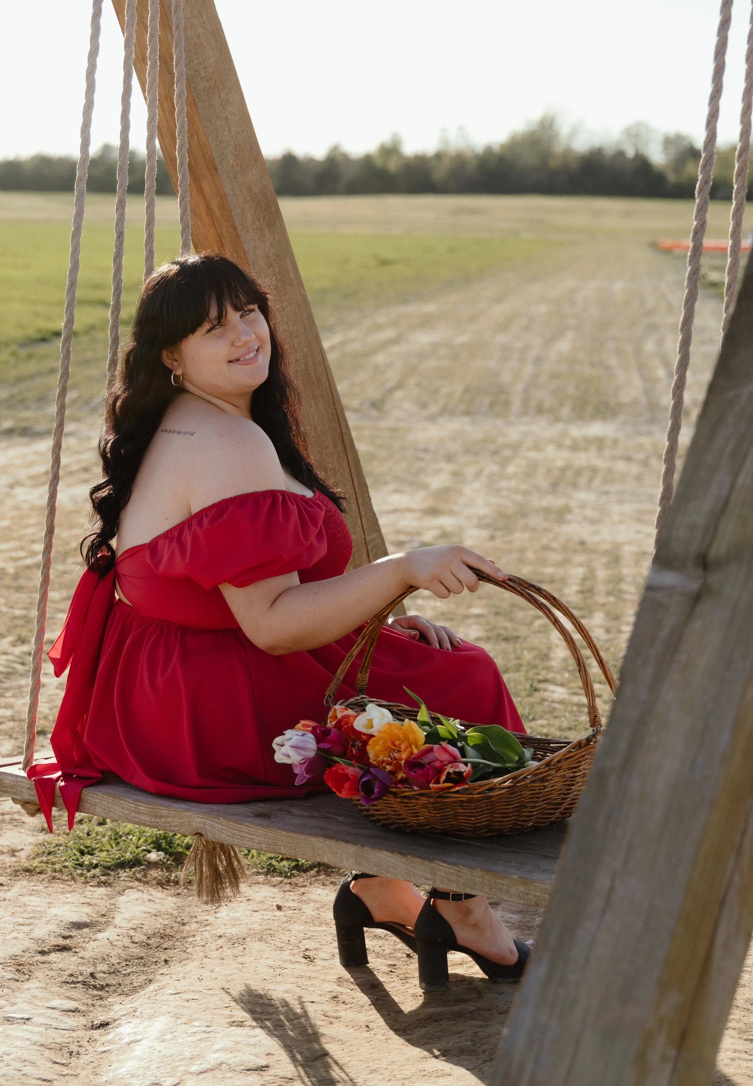 A woman in a red dress sitting on a wooden swing in an outdoor setting, holding a basket of colorful flowers, smiling, with a field and trees in the background.