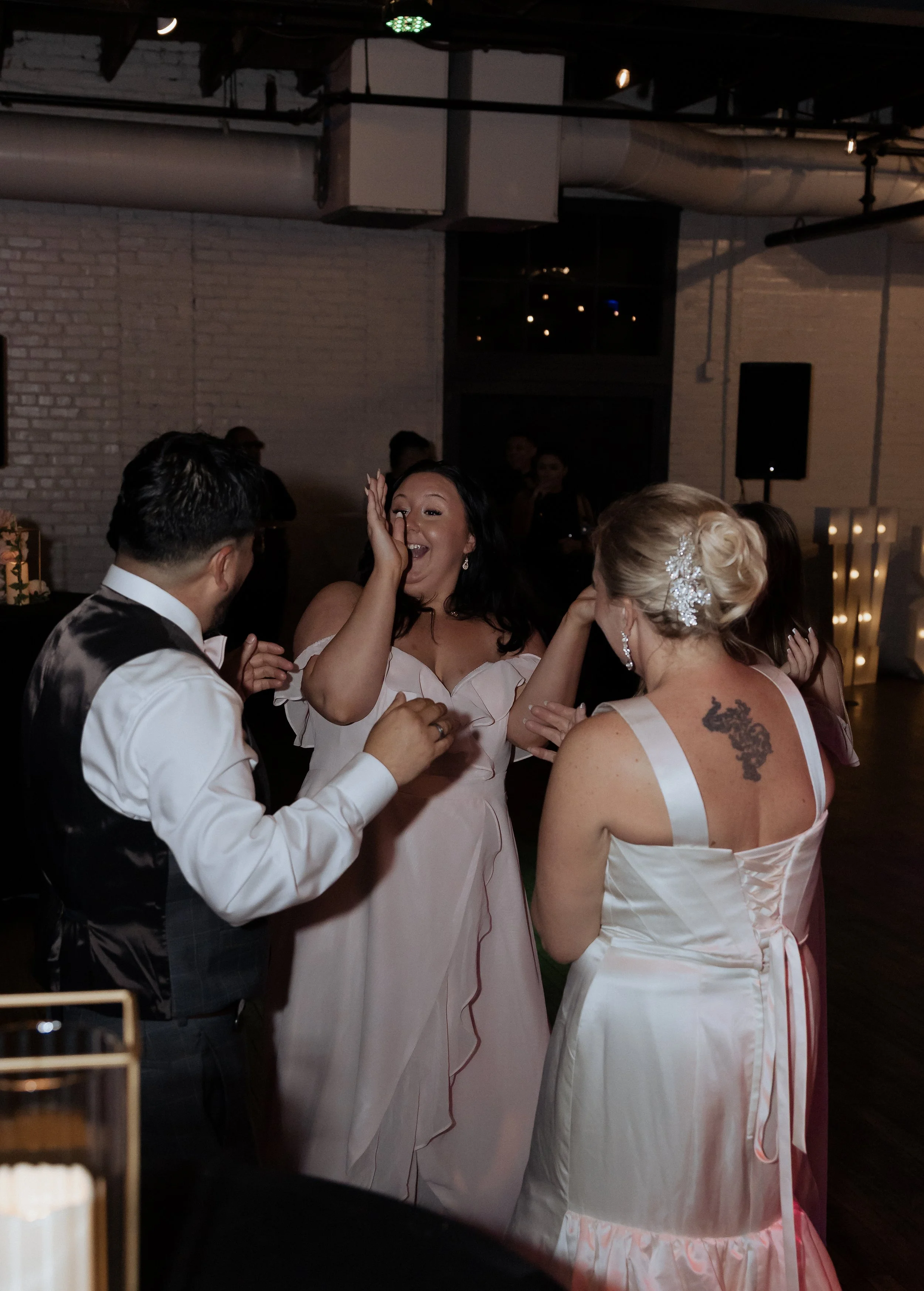 Group of people at a wedding reception, dancing and celebrating indoors, with a brick wall and dim lighting in the background.