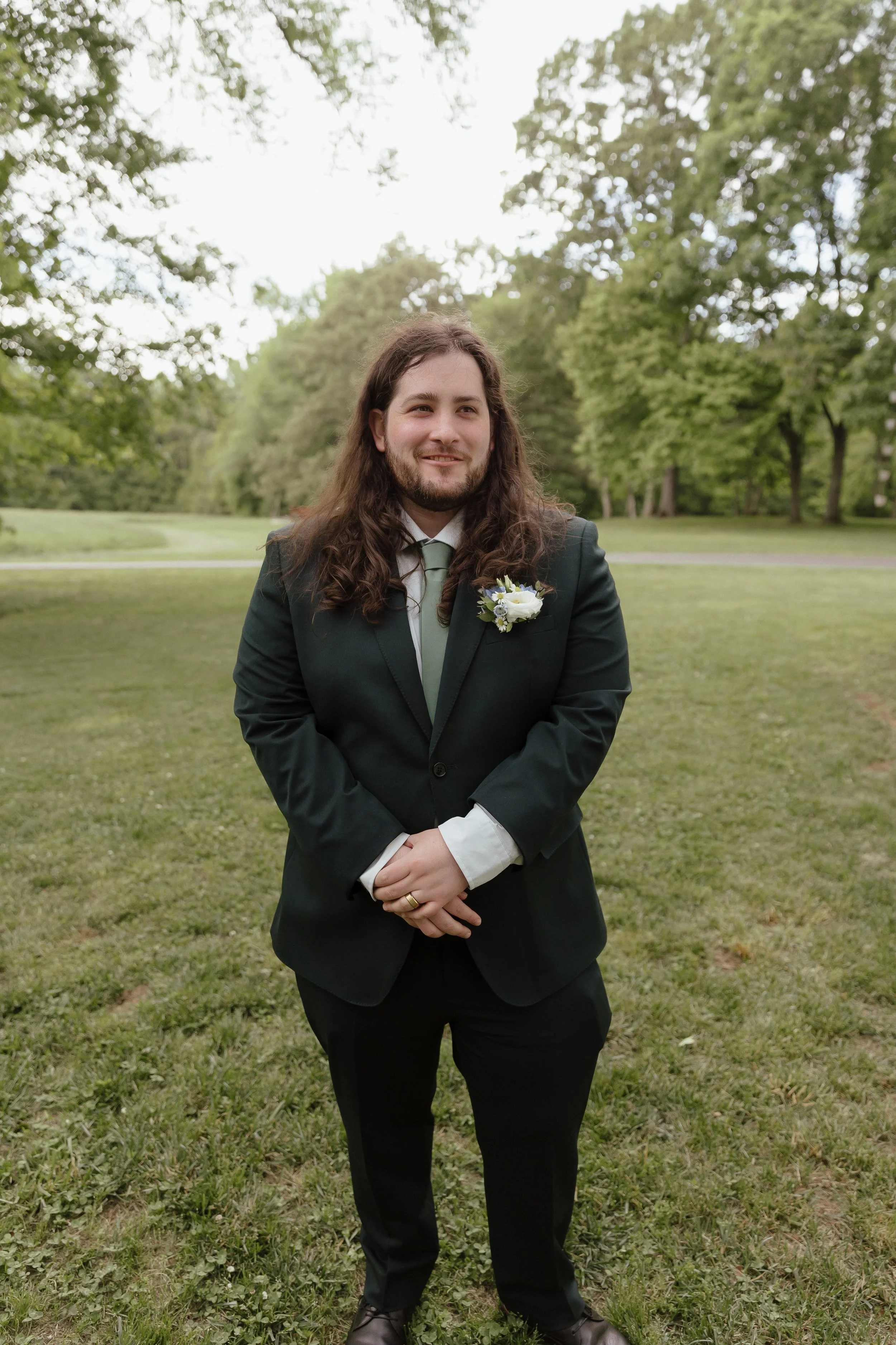 A photo of a man with long hair, standing in a dark green suit, smiling, with a floral boutonnière and sage green tie.