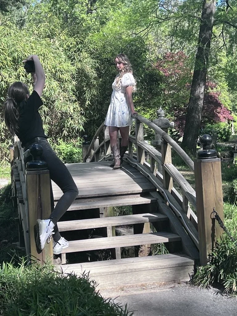A woman in a white dress and cowboy boots standing on a small arched wooden bridge, with another woman in black standing on the side of the bridge taking a photo of her, in a lush green garden setting.