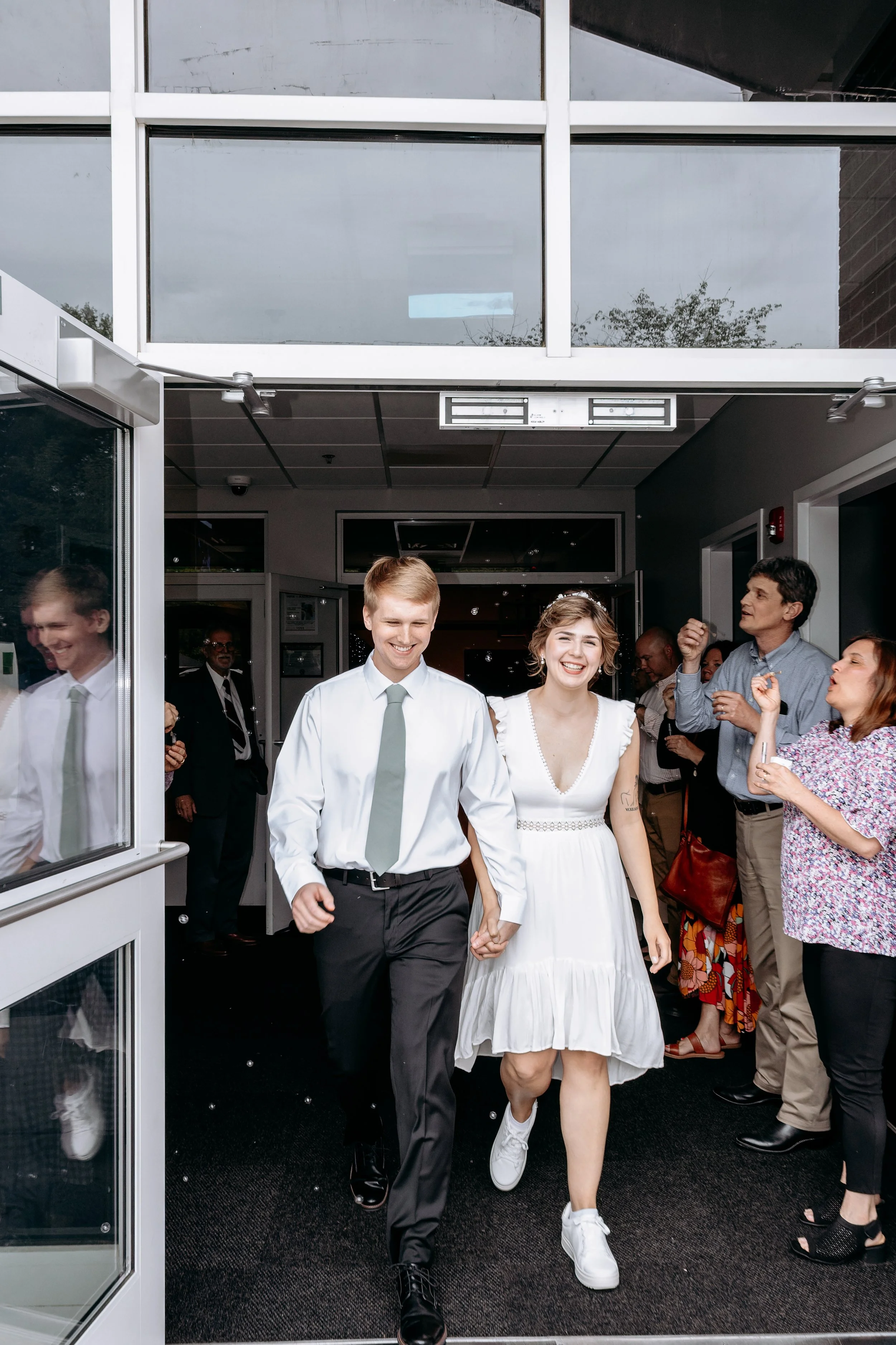 A young couple walking out of a building holding hands, smiling, with a group of people clapping and cheering behind them.