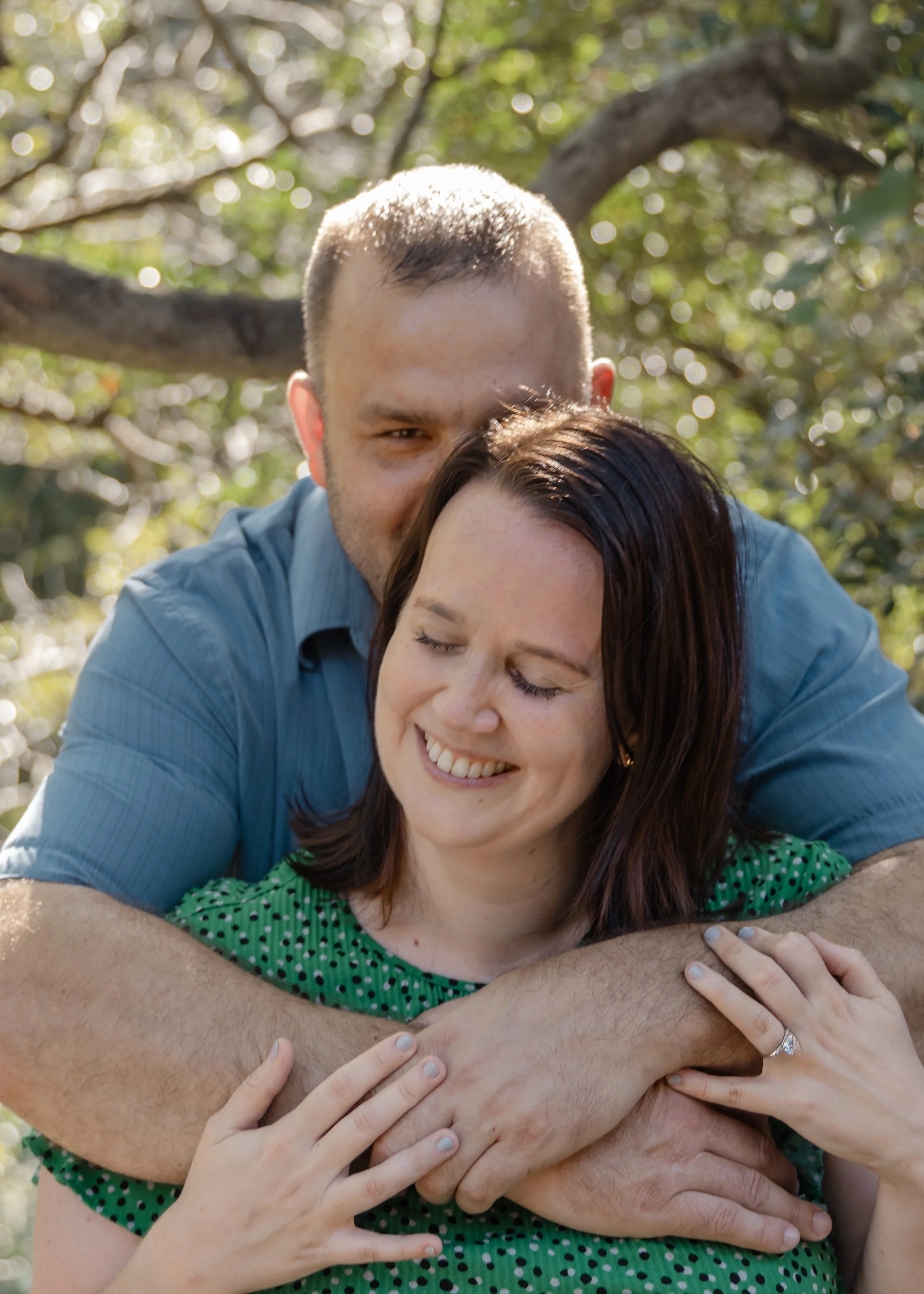 A man hugging a woman from behind outdoors, both smiling with eyes closed amidst sunlight and trees.