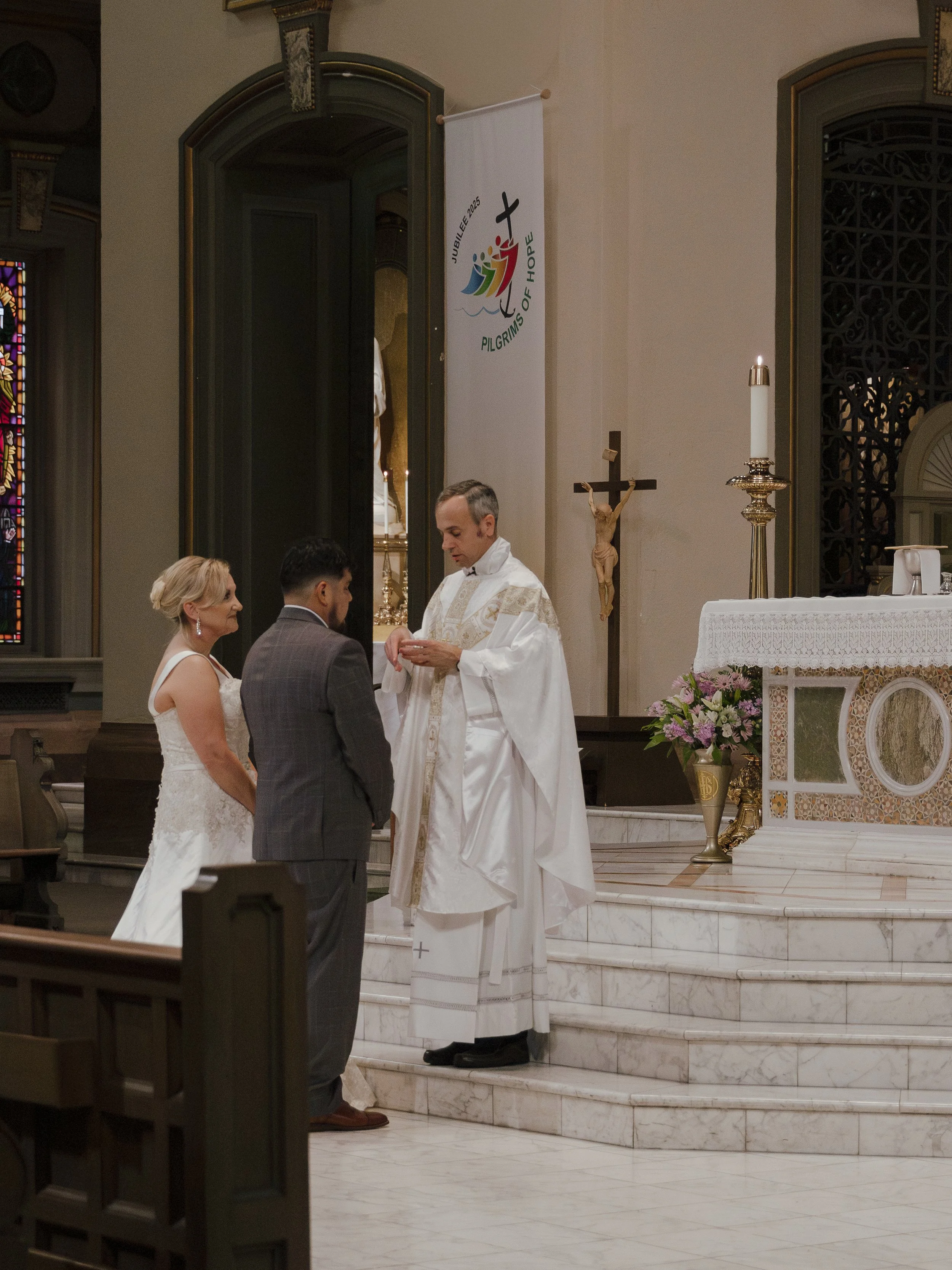 A wedding ceremony in a church, with a priest conducting the vows for a couple standing on the altar. The bride has blonde hair in an updo and is wearing a white wedding dress. The groom has dark hair and a beard, wearing a gray plaid suit. The churc