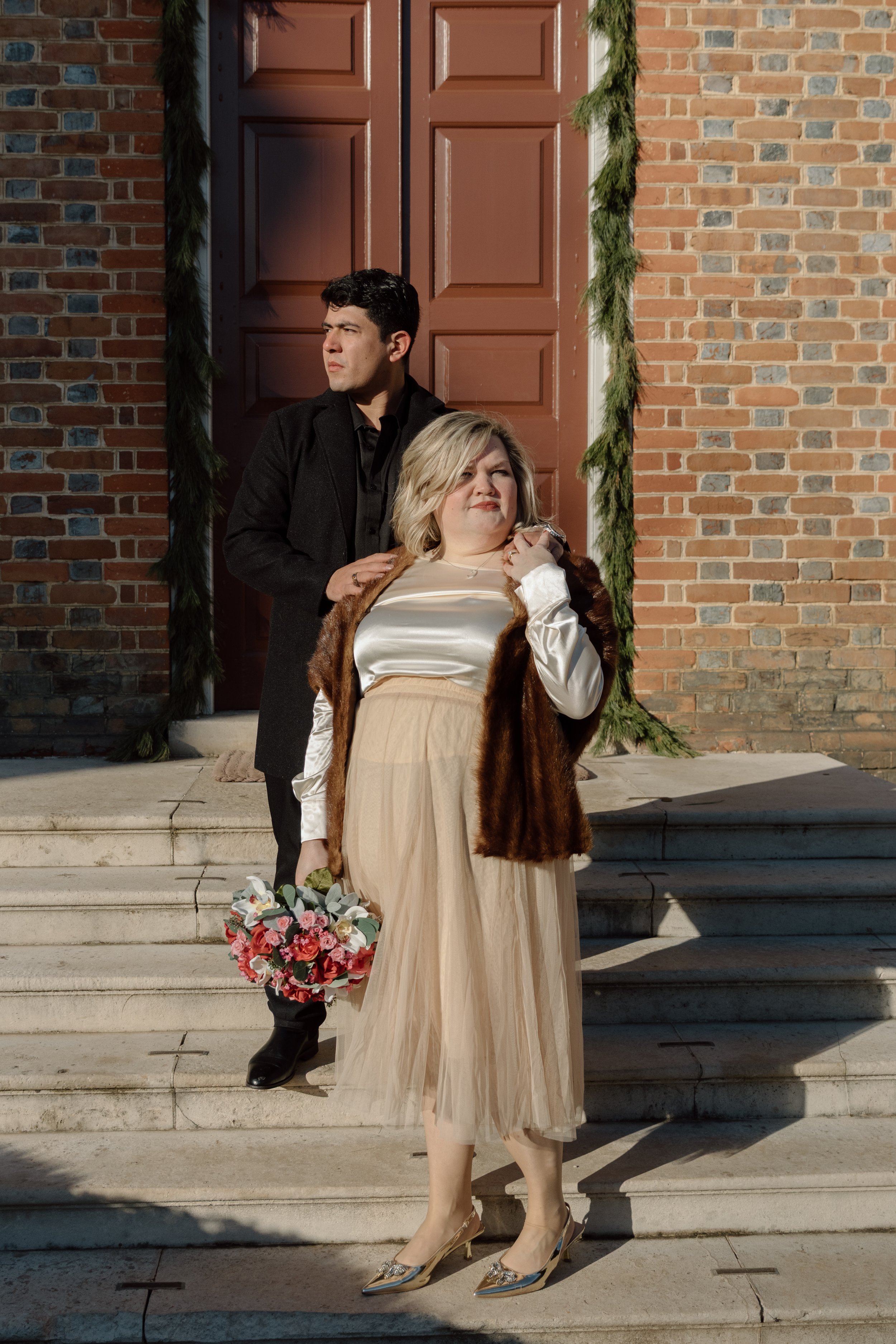 A woman with blonde hair wearing a beige tulle skirt, a white satin blouse, and a fur coat, holding a bouquet of pink and white flowers, standing on church steps. A man with dark hair in a black coat stands behind her, with one hand on her shoulder, 