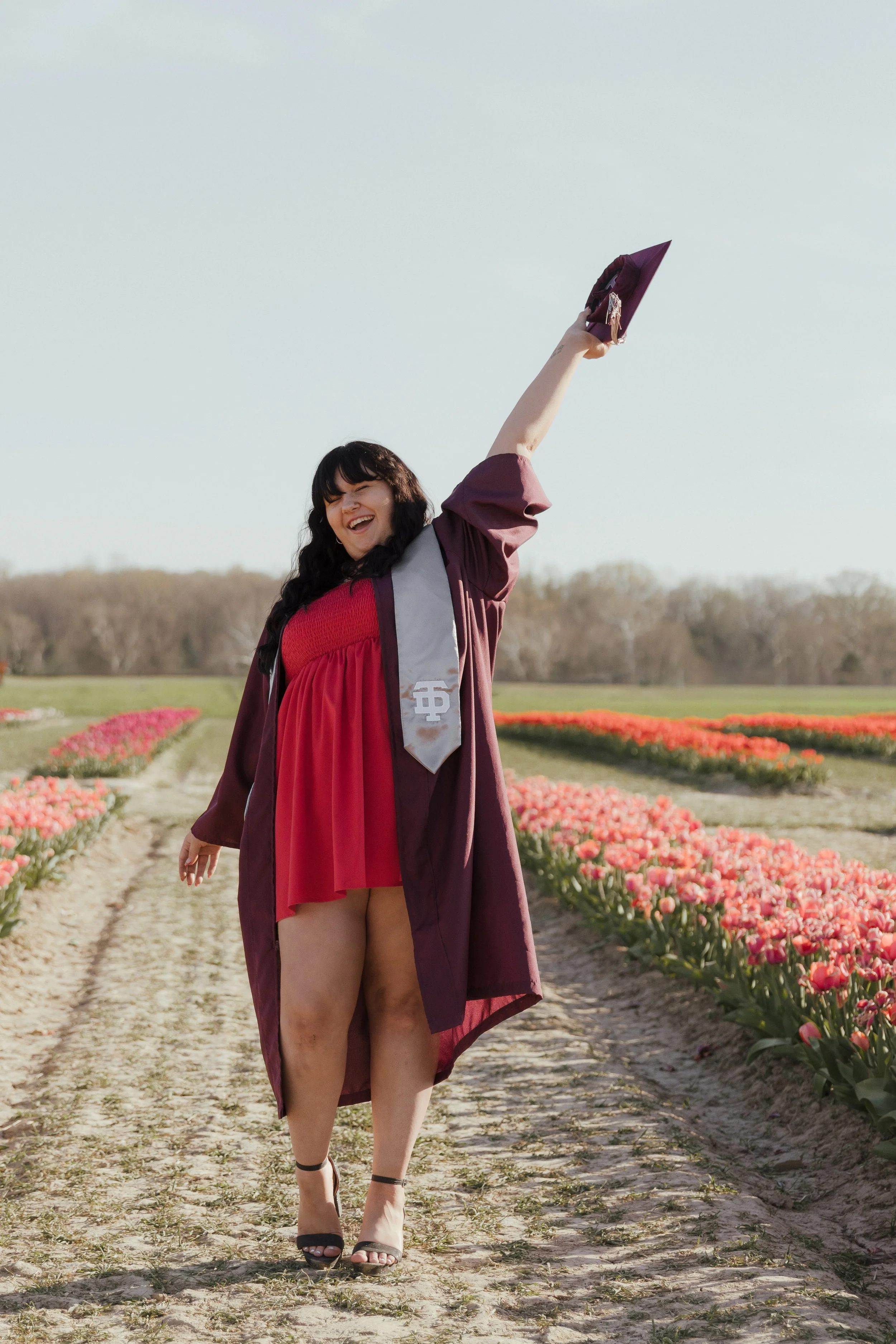 A young woman dressed in a red dress and graduation gown holding her cap high and celebrating in a field of pink tulips.
