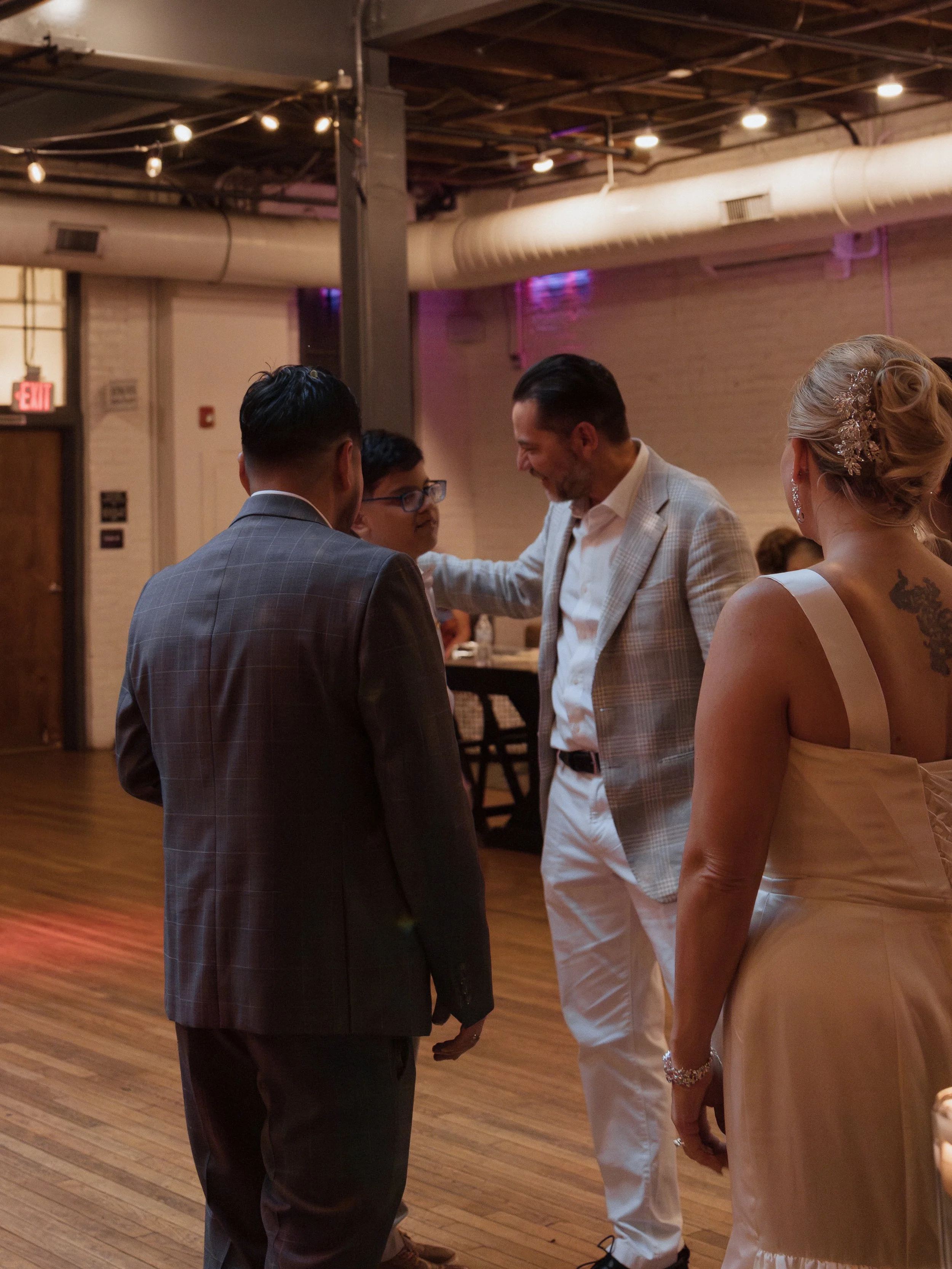 A group of people dancing at a wedding reception in a rustic indoor venue with wood floors and exposed brick walls.