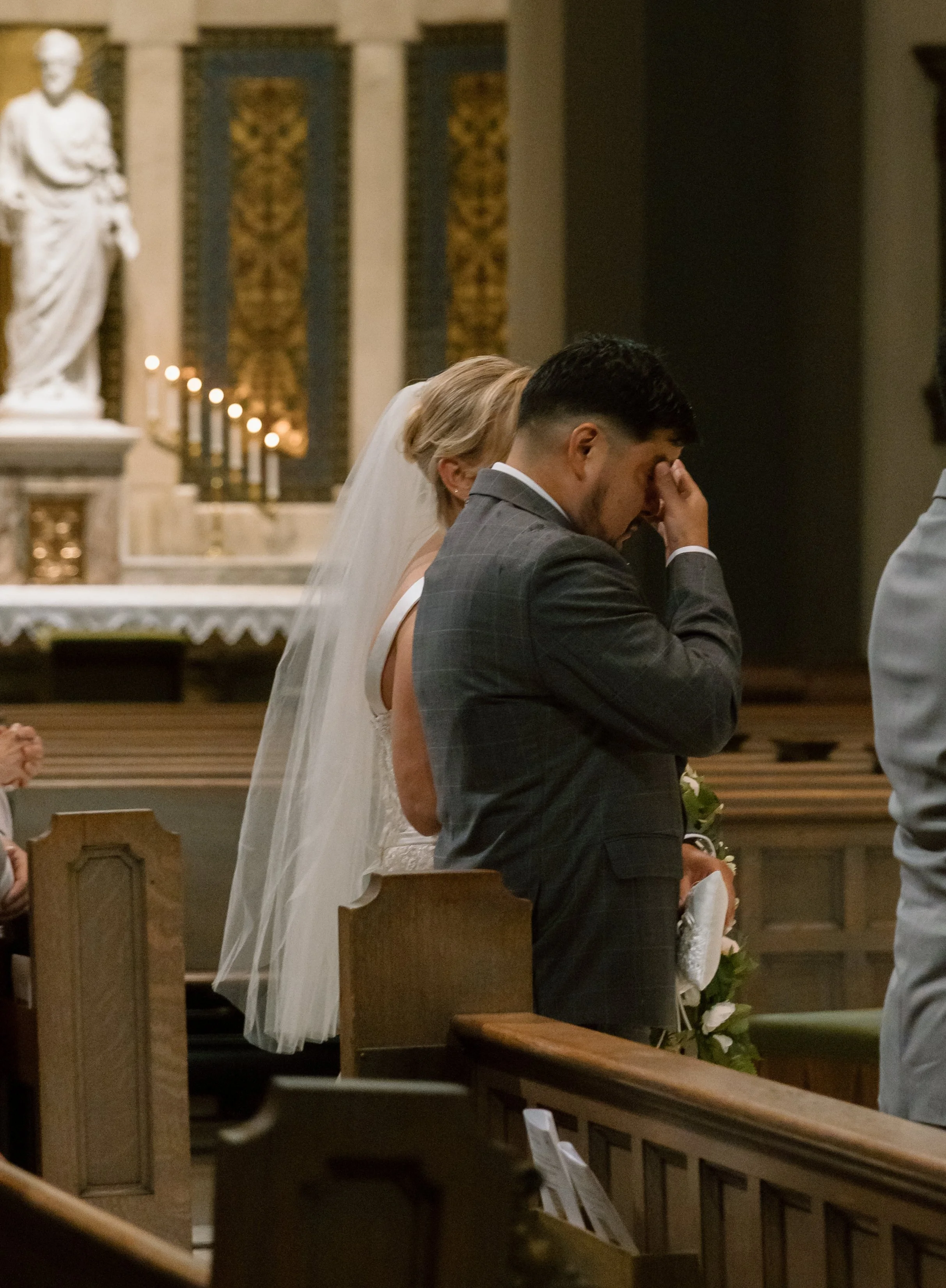 A bride and groom kneeling in prayer during a wedding ceremony inside a church, with a statue and candles in the background.