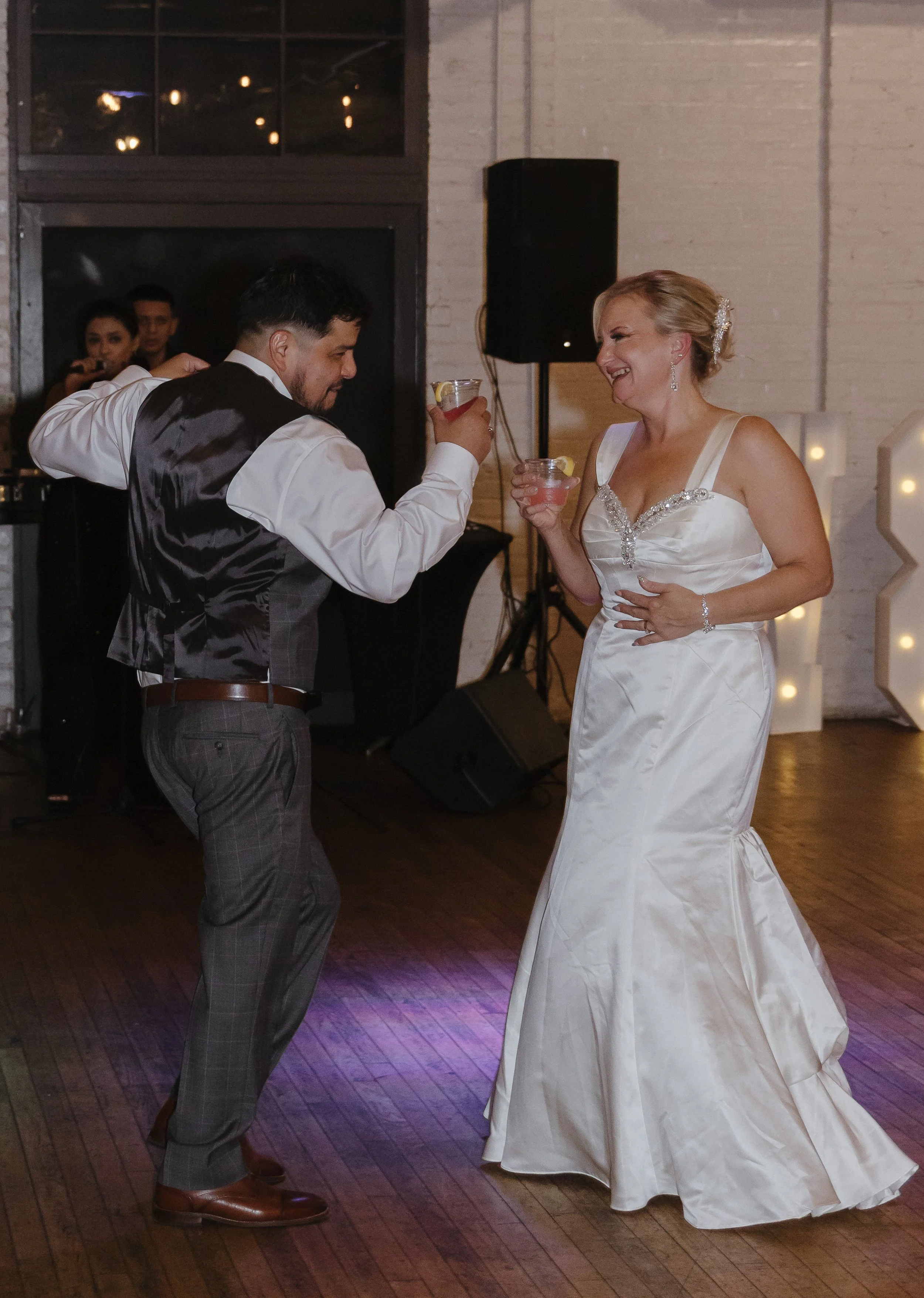 A bride and groom dancing at their wedding reception, holding cocktails and smiling at each other.