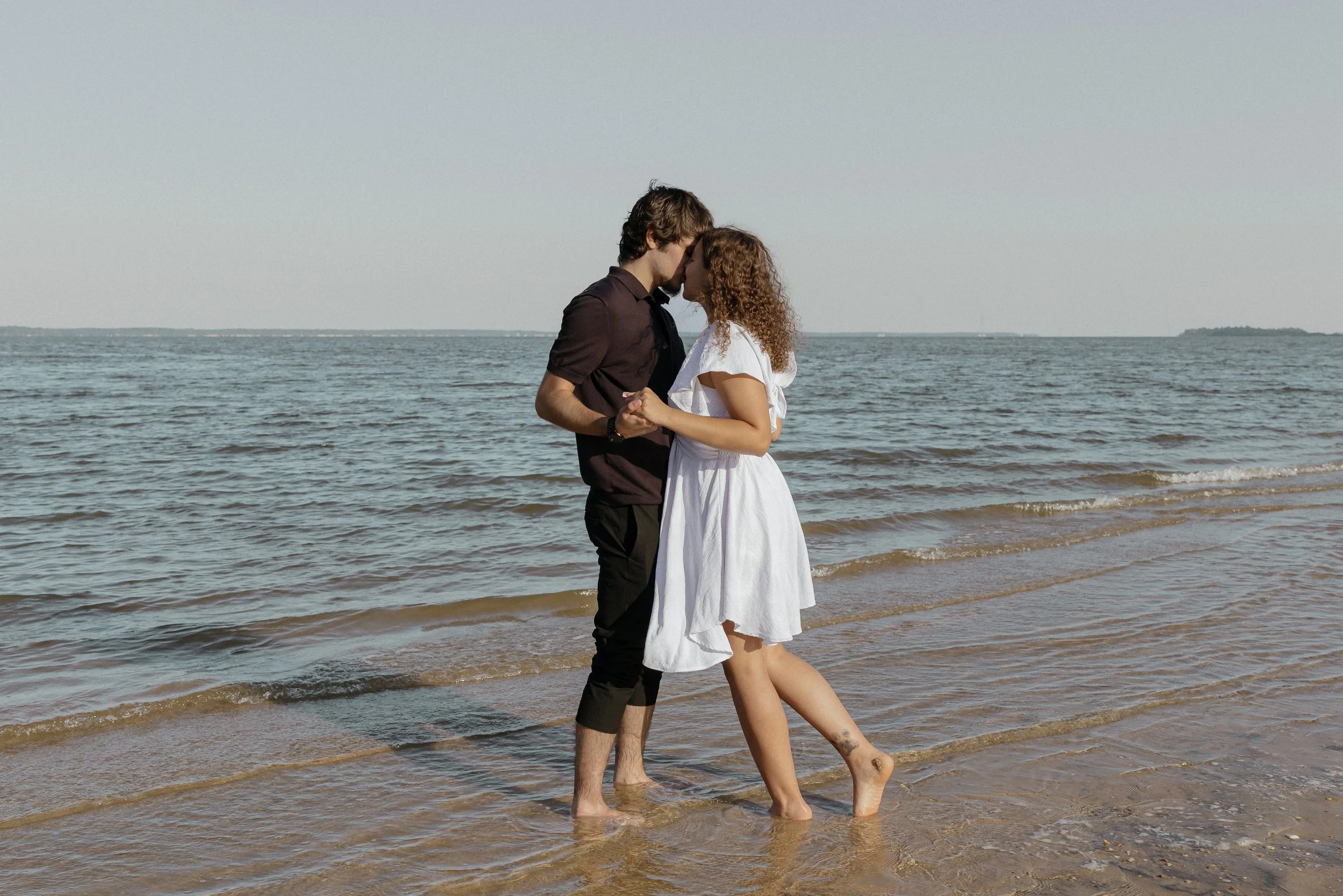A couple holding hands and leaning in for a kiss at the beach, standing in shallow water with the ocean and sky in the background.
