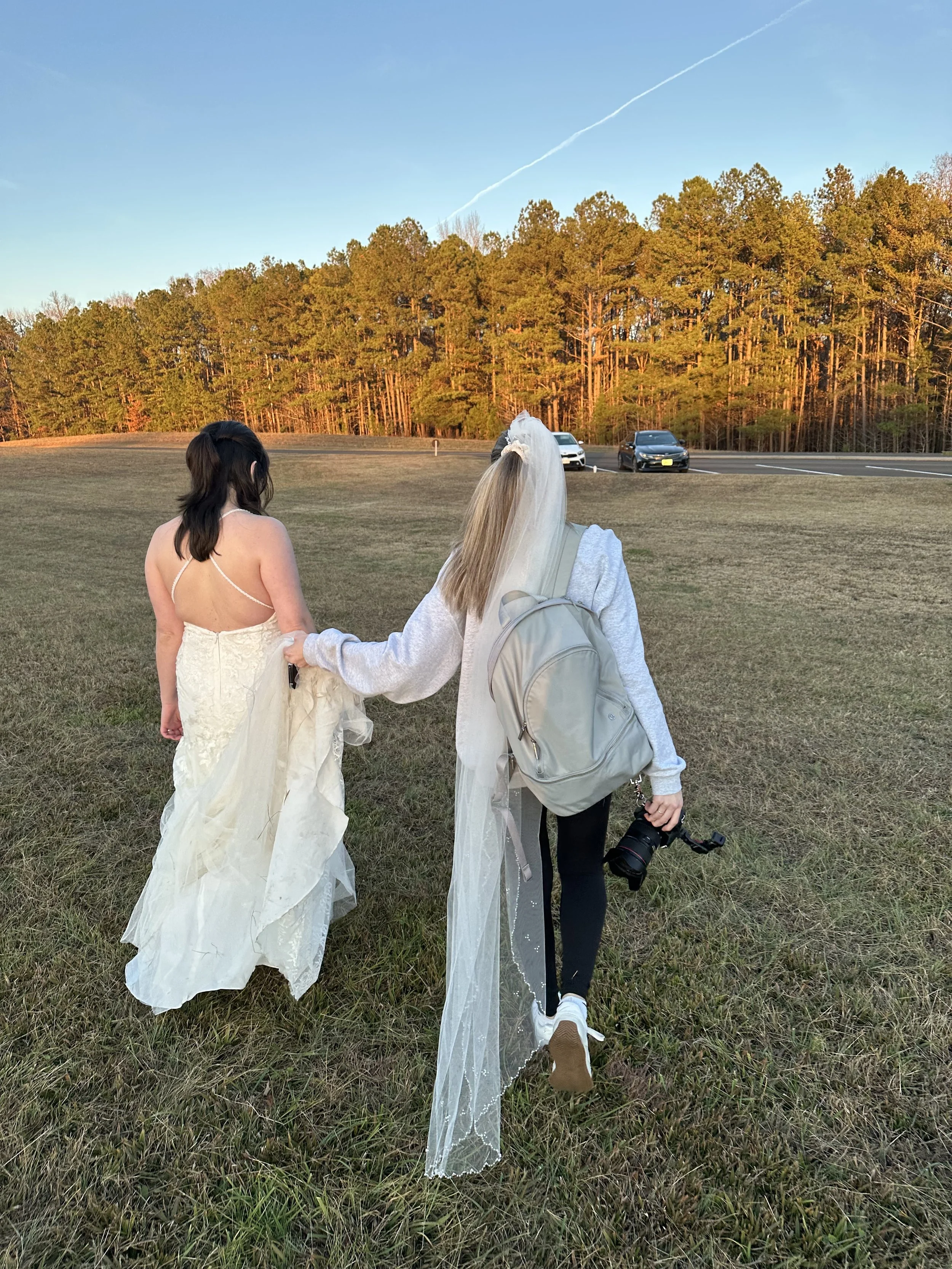 A bride in a wedding dress walking on a grassy field with a woman carrying a camera, holding her hand.