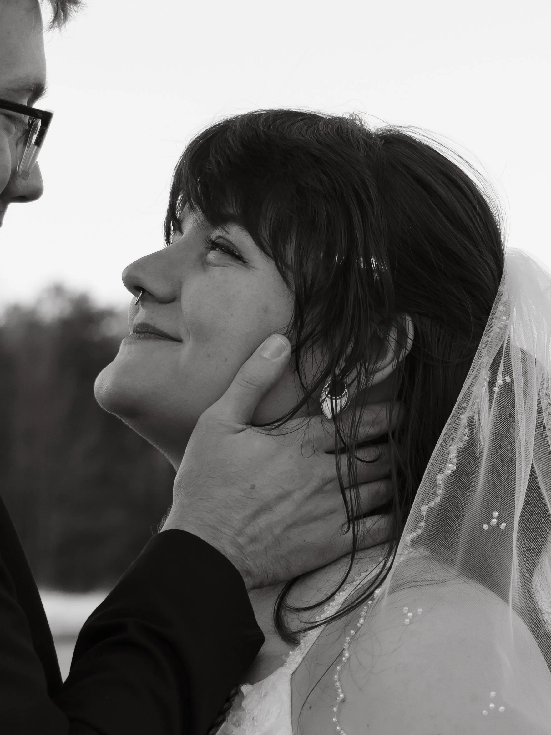 A couple on their wedding day, with the groom gently holding the bride's face, both smiling happily, outdoors in black and white.