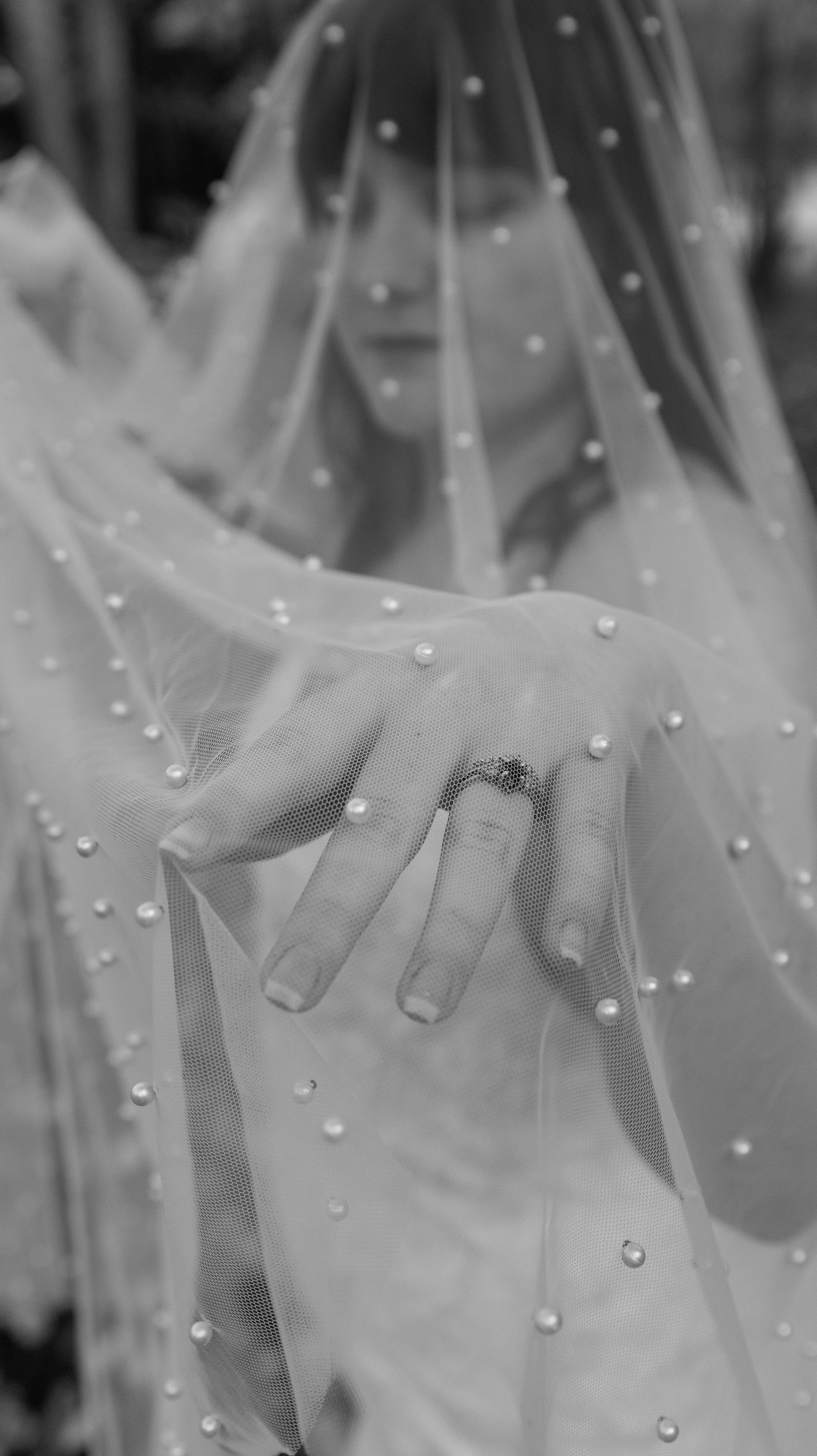 A person behind a sheer veil decorated with small pearls, touching the veil with their hand, with their face partially visible through the fabric.