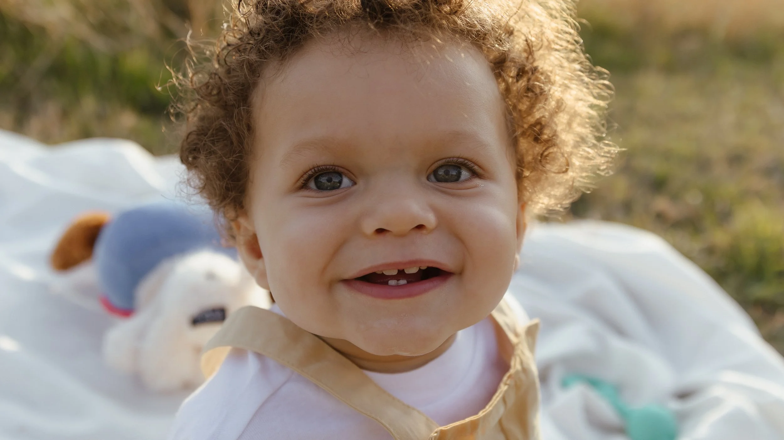 Close-up of a young child with curly hair smiling outdoors, with a plush toy dog in the background.