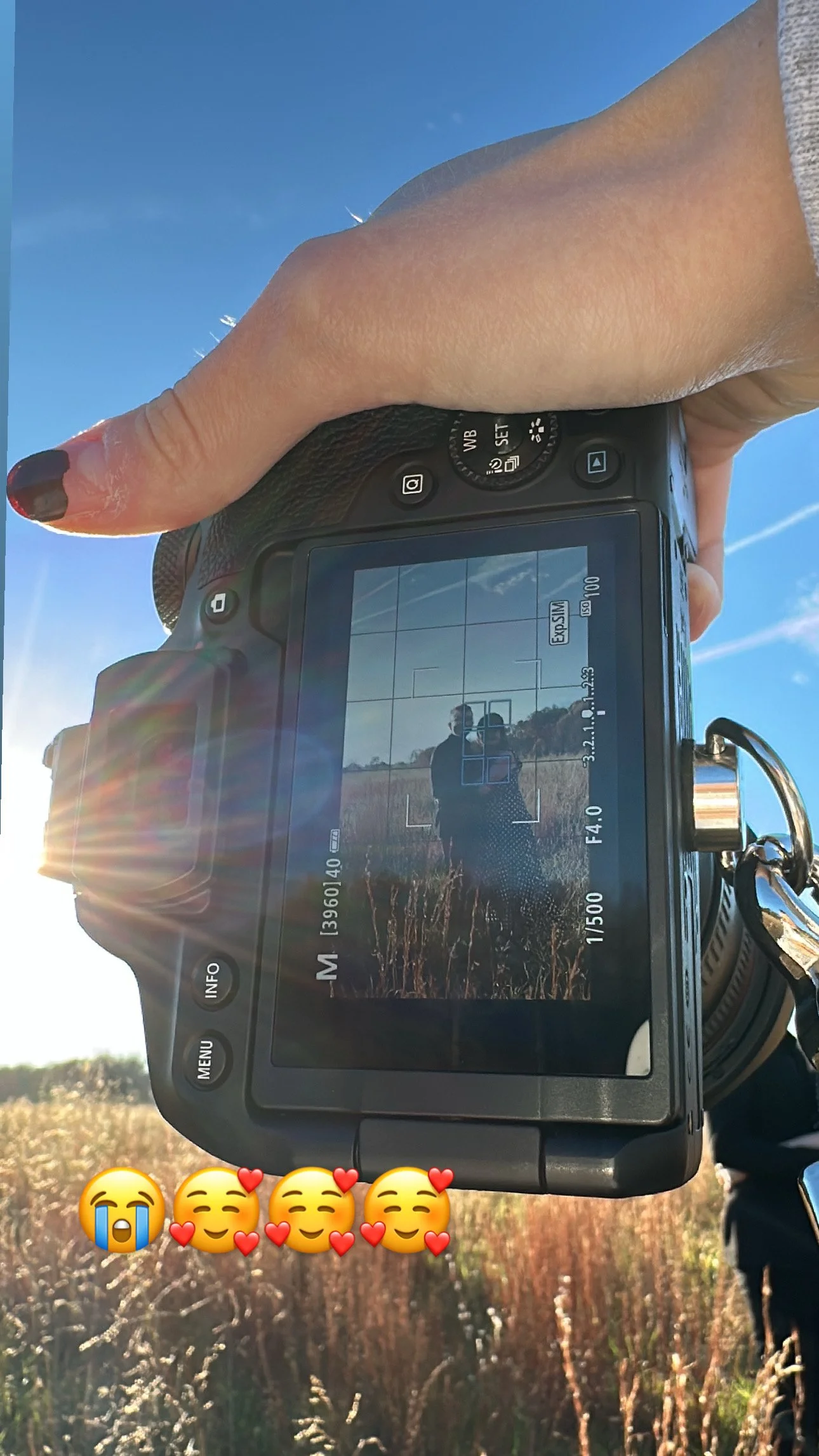 A person holding a camera outdoors during daytime, capturing a photo of themselves and another person in a field with tall grass under a clear blue sky. The person's thumb is visible, and they have painted nails. Sunlight creates a lens flare in the image.