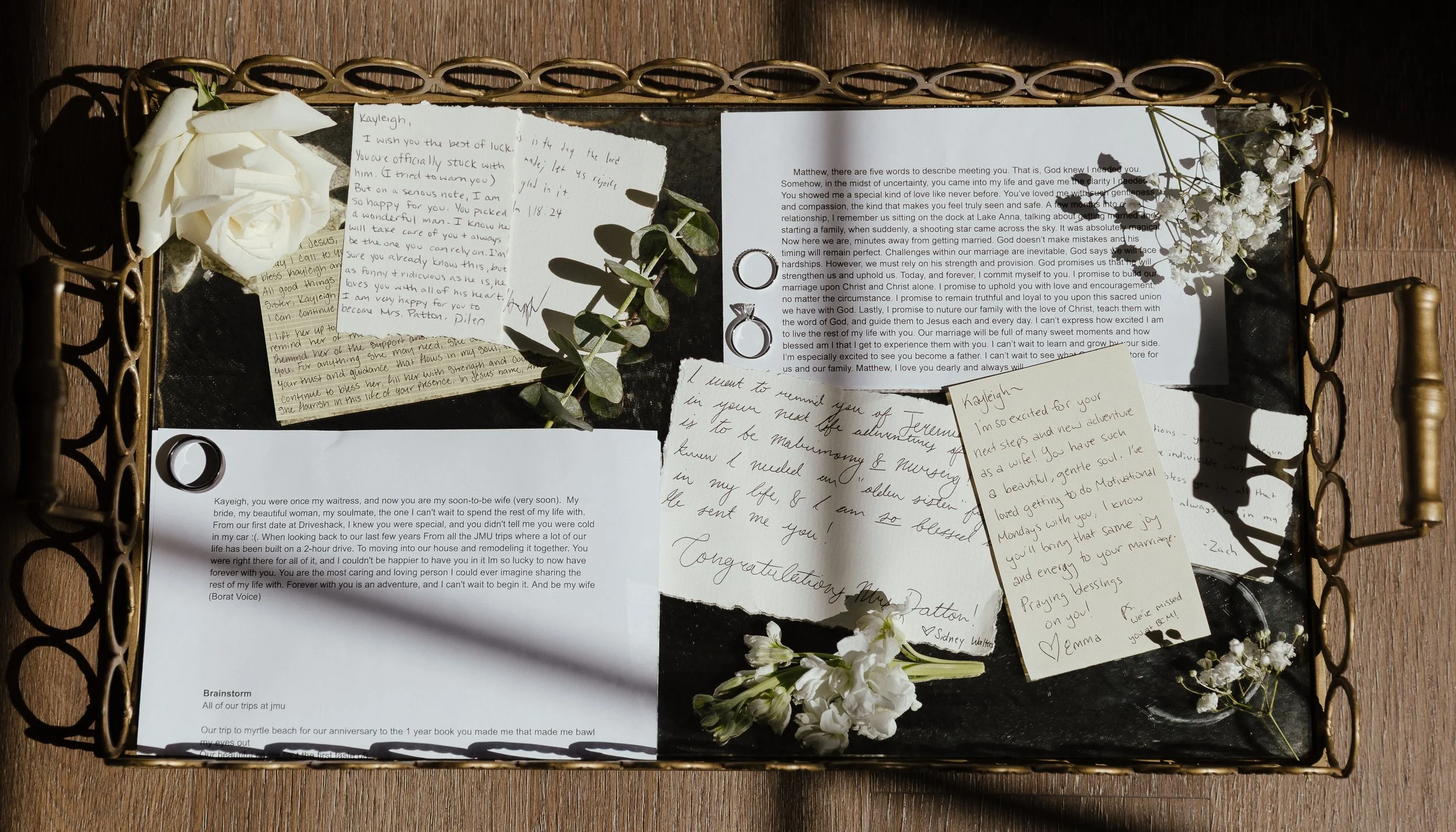 A detail photo of letters to the bride and the bride and groom's first touch notes laying on a golden mirror tray. The photo features loose florals like babies breath and a rose, and the wedding rings for the couple, taken in the light of a window.