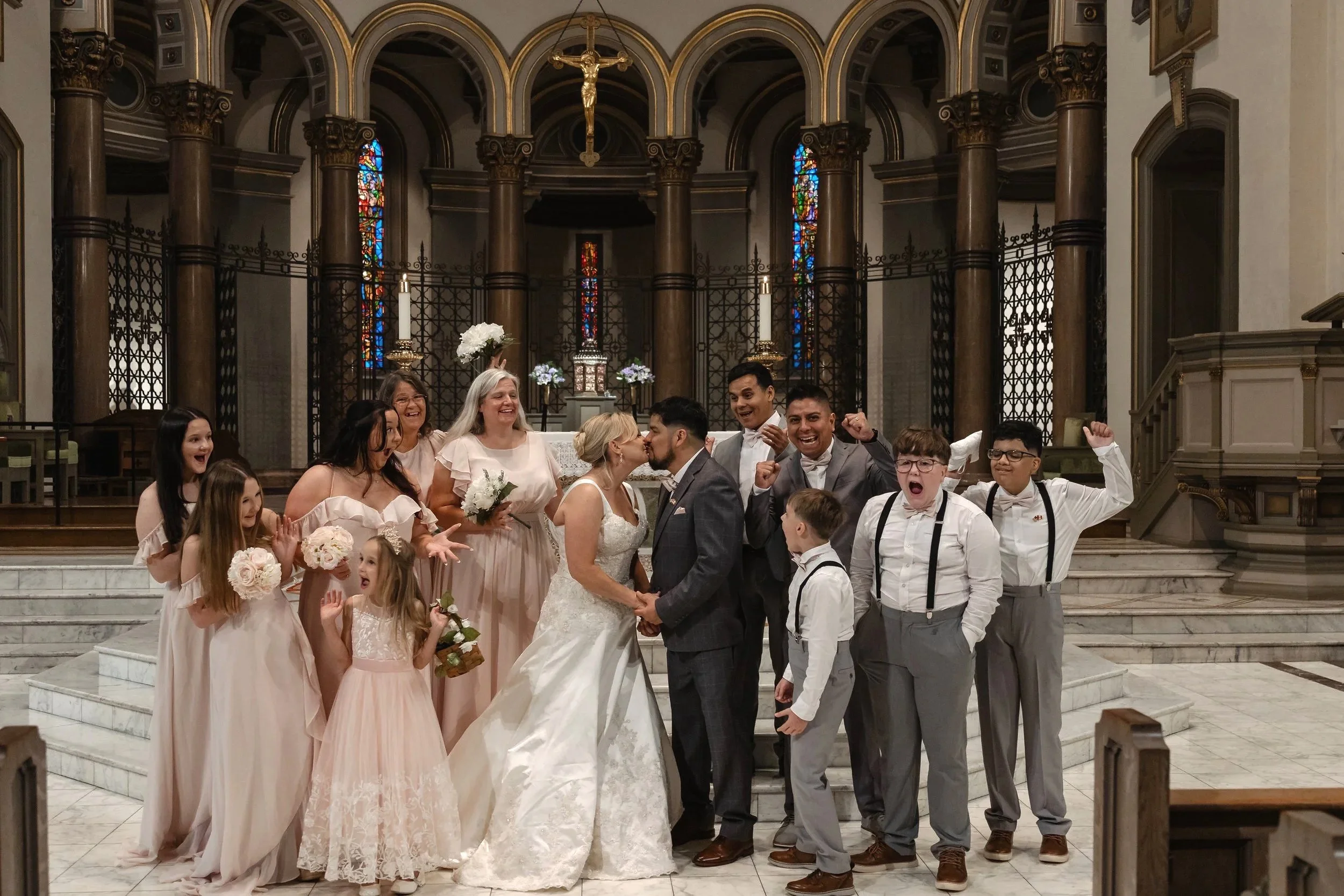 A group of people celebrating a wedding inside a church with stained glass windows and religious decor. The bride and groom are kissing in the center, surrounded by family and friends cheering and smiling.