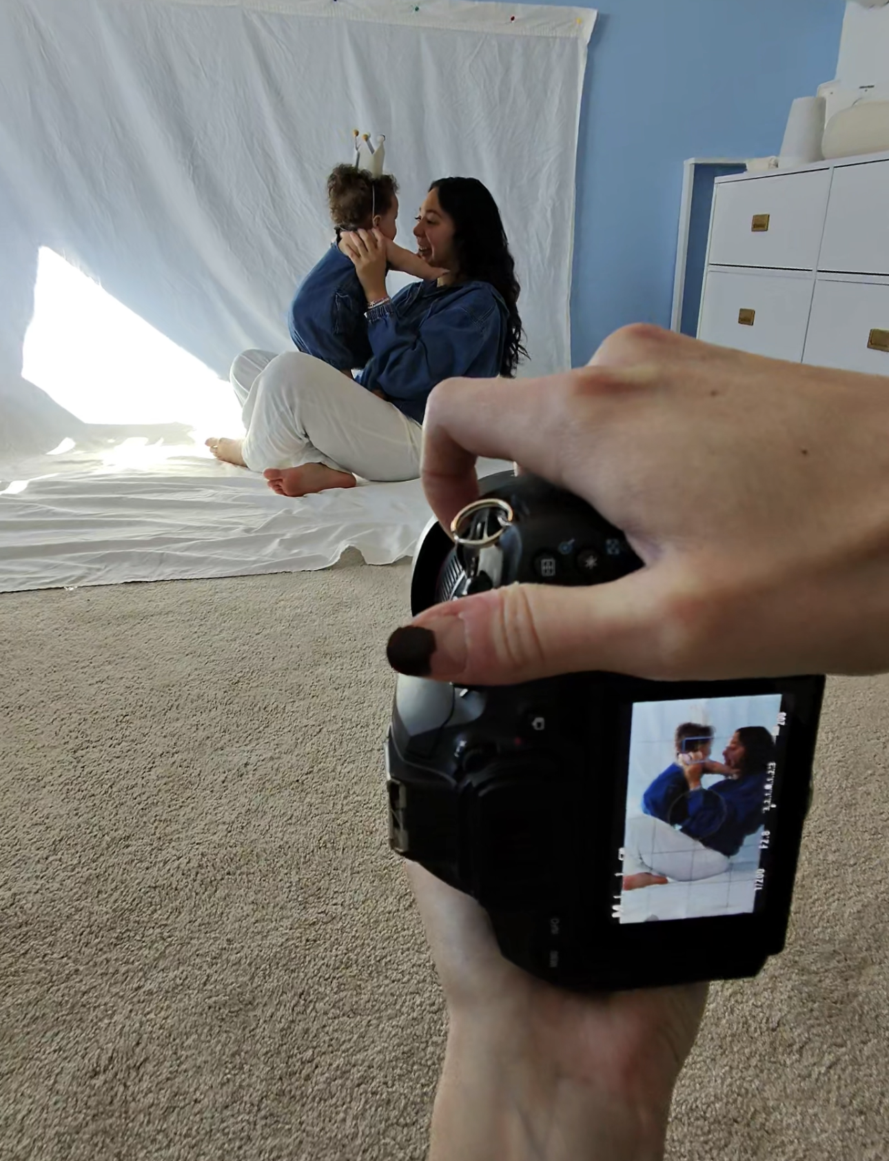 A woman sitting on a white sheet with a young girl, both smiling and touching faces, in a room with blue walls, white furniture, and a white sheet backdrop; a person taking a photo with a camera in the foreground.