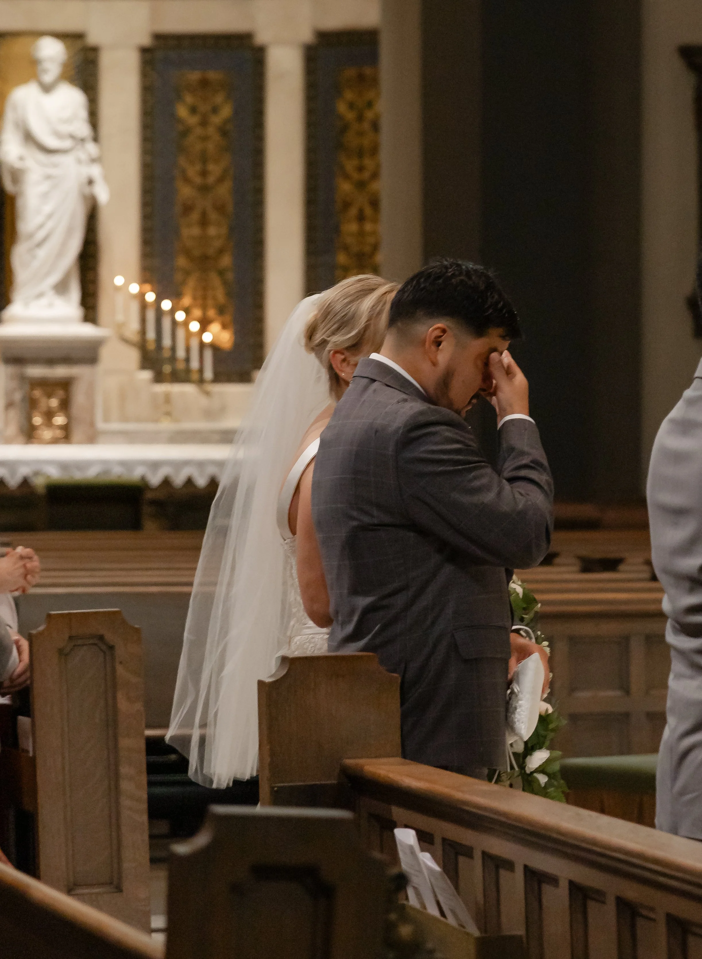 A bride and groom kneeling during a wedding ceremony in a church, with the groom pinching the bridge of his nose and the bride partially obscured behind him, in front of an altar and a statue.