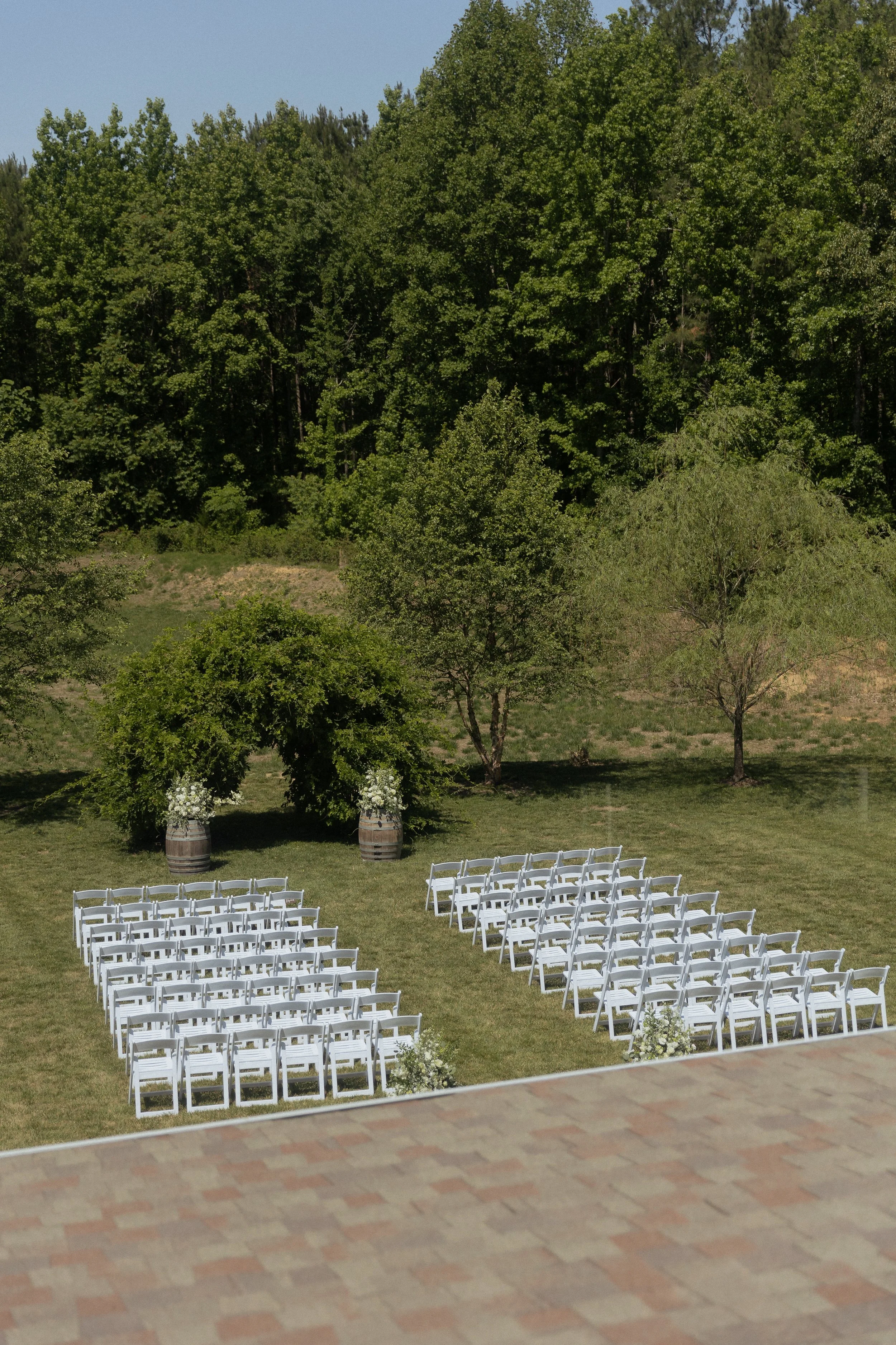 A photo of a wedding ceremony space from the view of a venue window. White chairs where the guests will sit are lined up facing a natural arbor of lush greenery, complimented with two adjacent barrels with white florals on top of them.