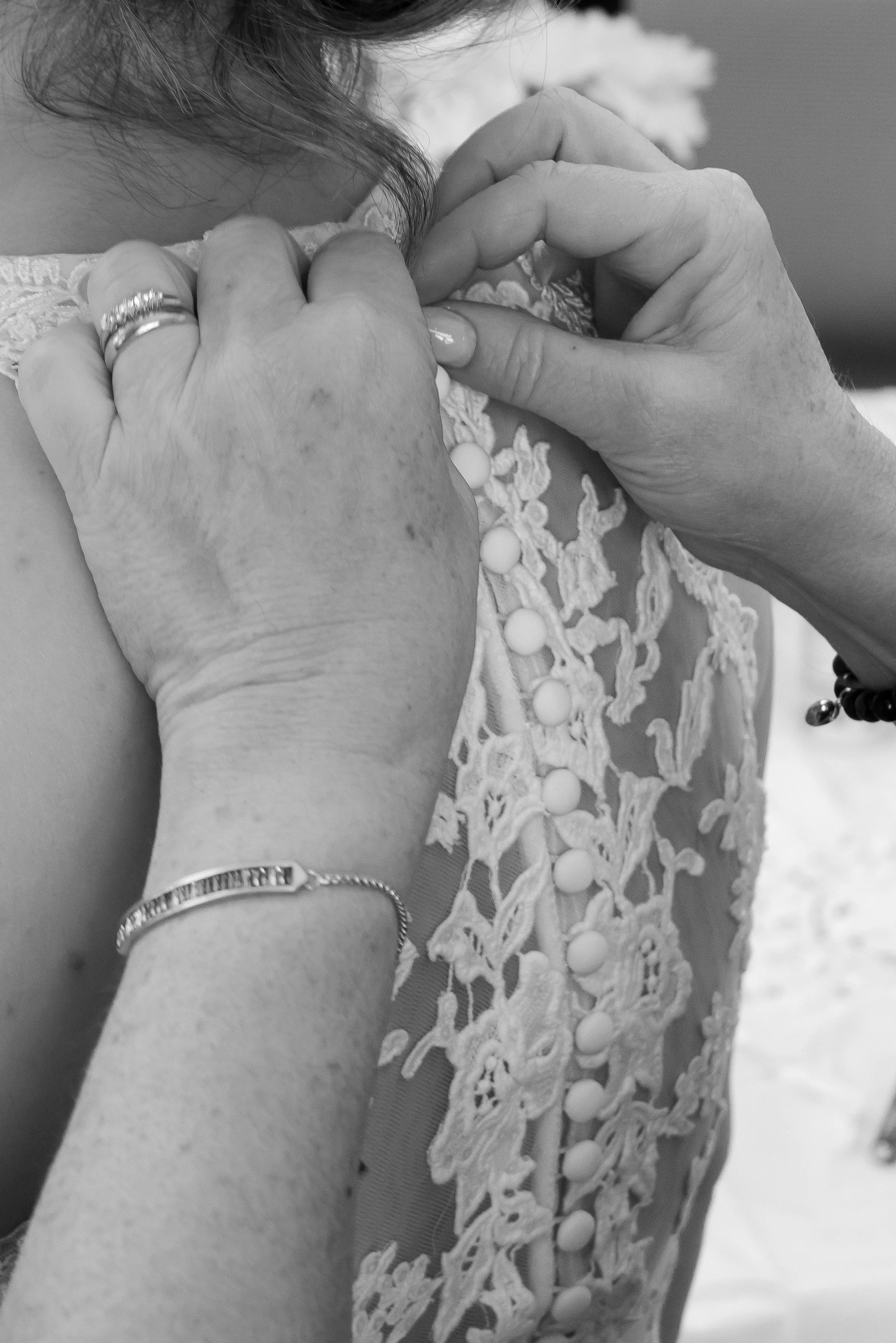 Close-up of an elderly woman pinning or adjusting a lace dress with buttons, focusing on her hands and part of her face, in black and white.