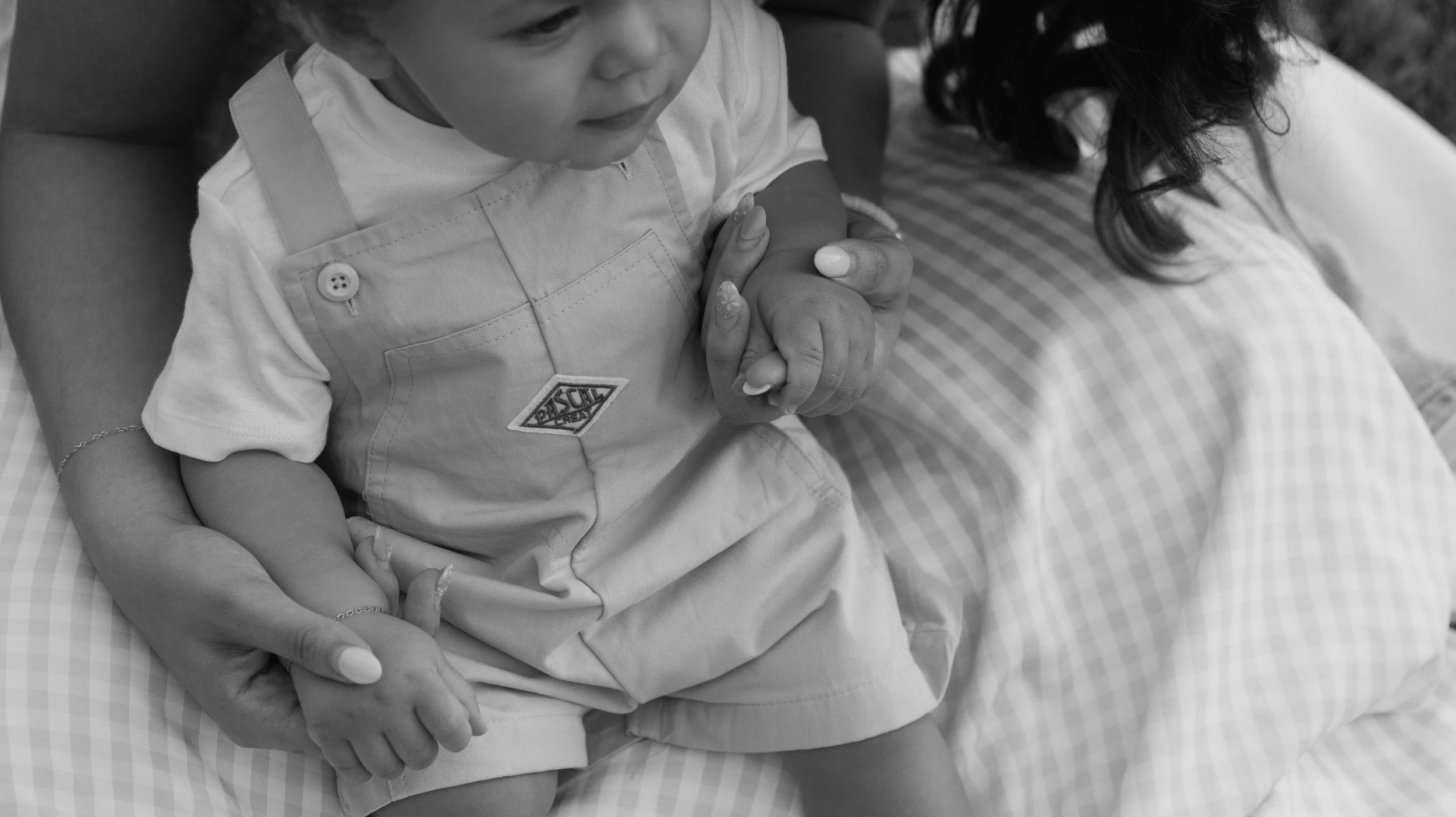 A child wearing a light-colored shirt and overalls sitting on someone's lap, holding hands with an adult. The scene appears to be outdoors on a checkered blanket.