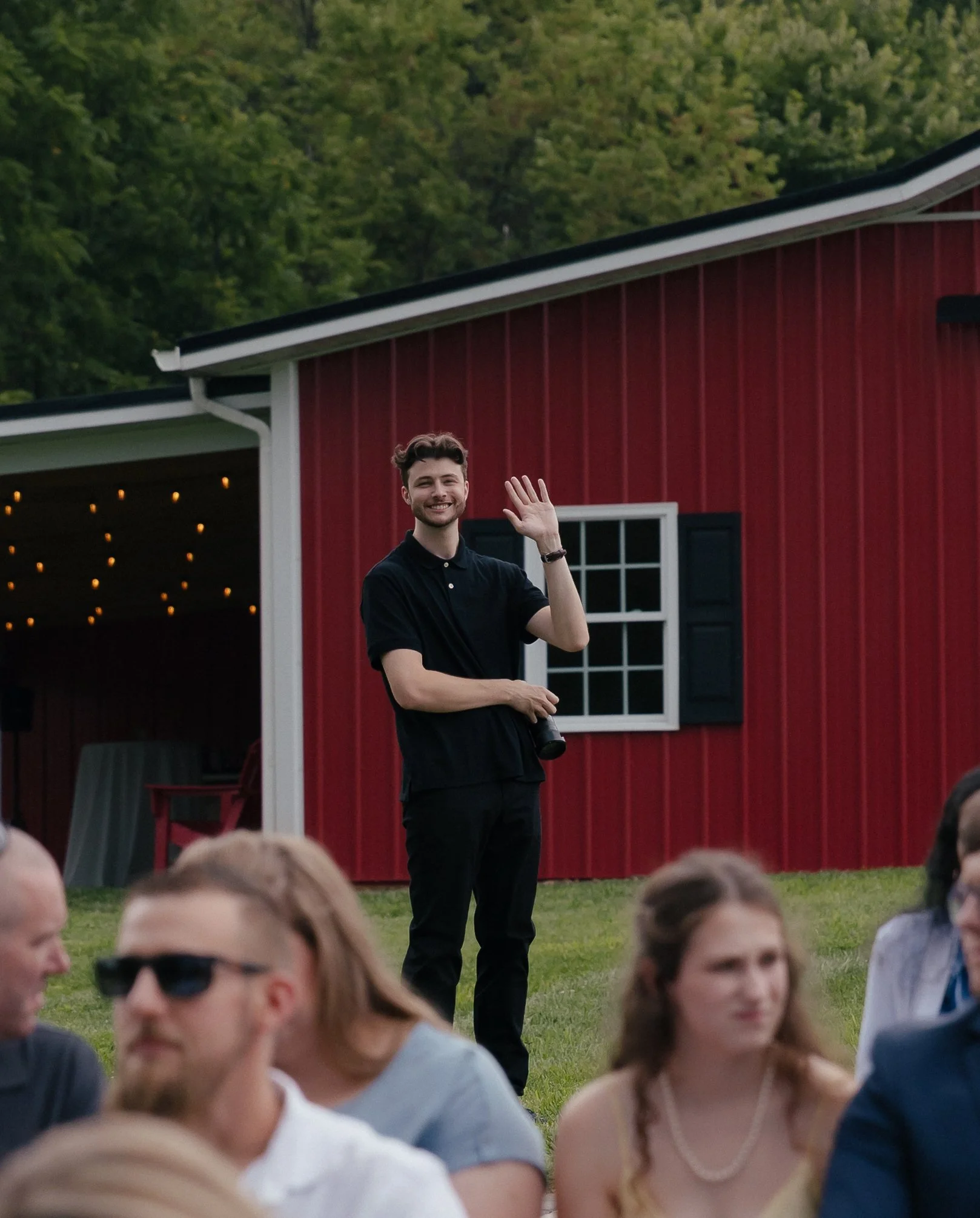 A man holding a camera in front of a red barn, standing behind wedding guests at a Virginia wedding venue.