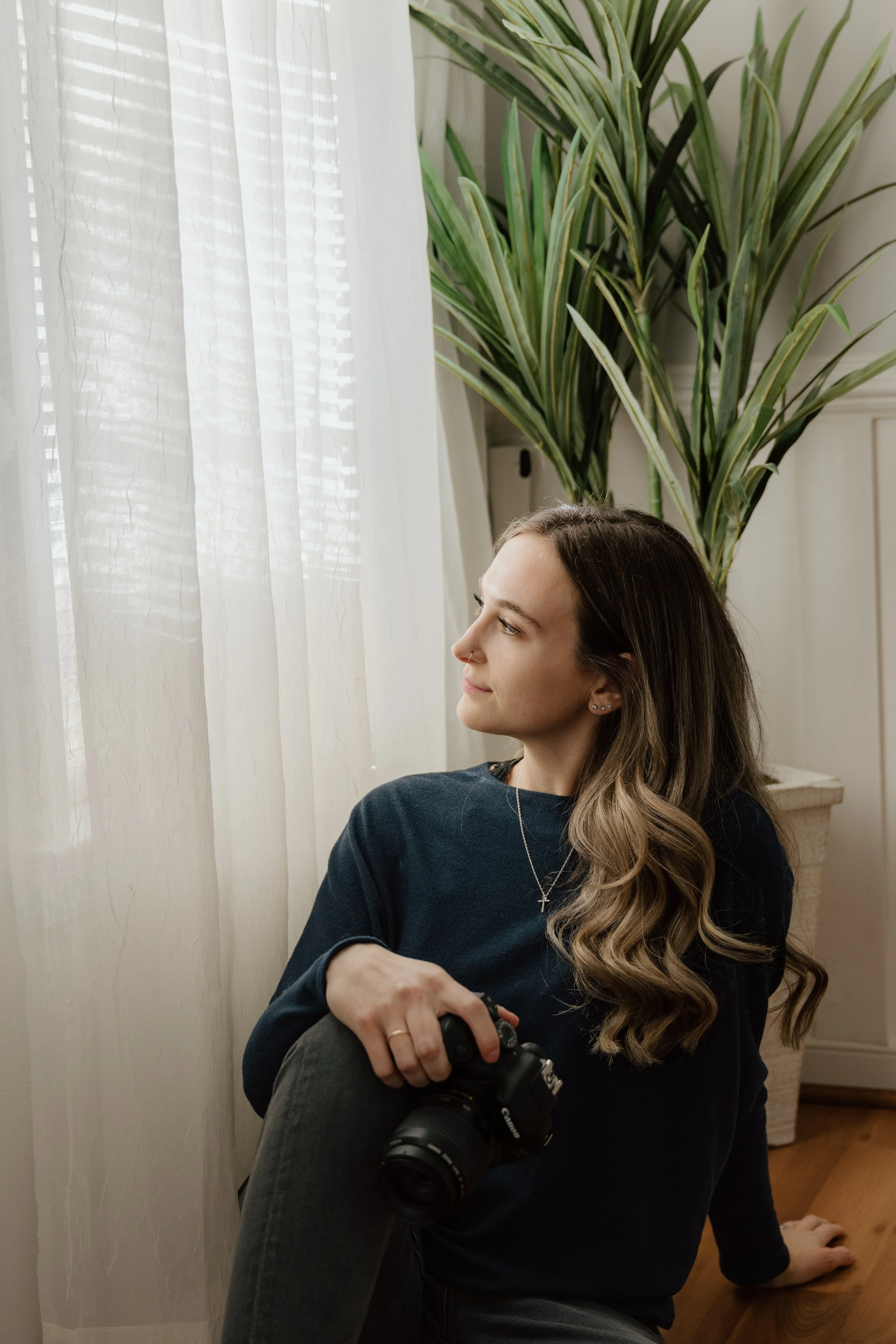 A young woman with long, wavy brown hair sitting on the floor next to a window with sheer white curtains, holding a camera in her right hand, and looking out the window. There is a tall green potted plant behind her.