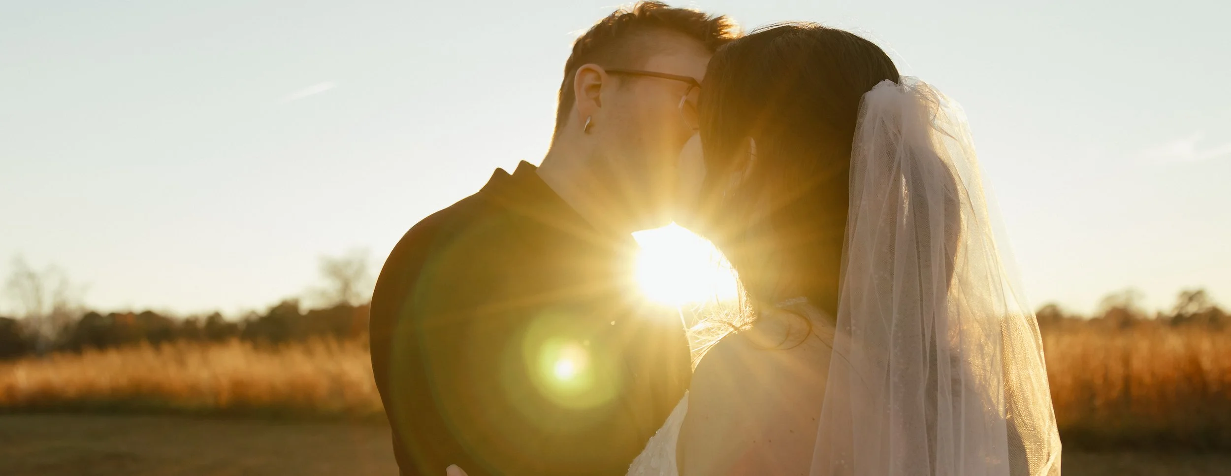 A couple sharing a kiss outdoors at sunset, with the sunlight shining between them and the bride wearing a veil.