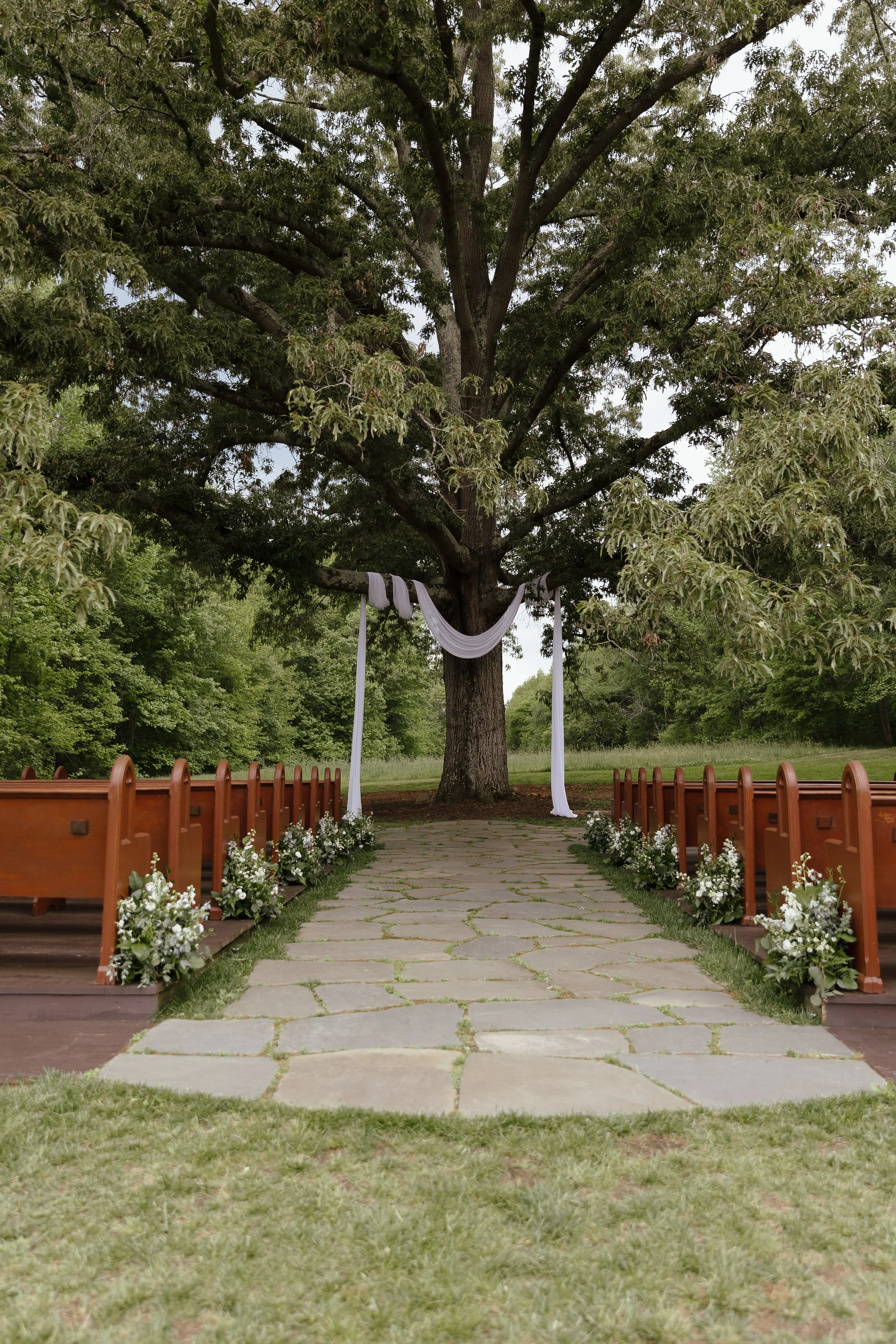 A photo of a wedding ceremony space with a white sheer cloth draped from a tree at the end of the aisle. The space features floral aisle decor, and wooden pews. 