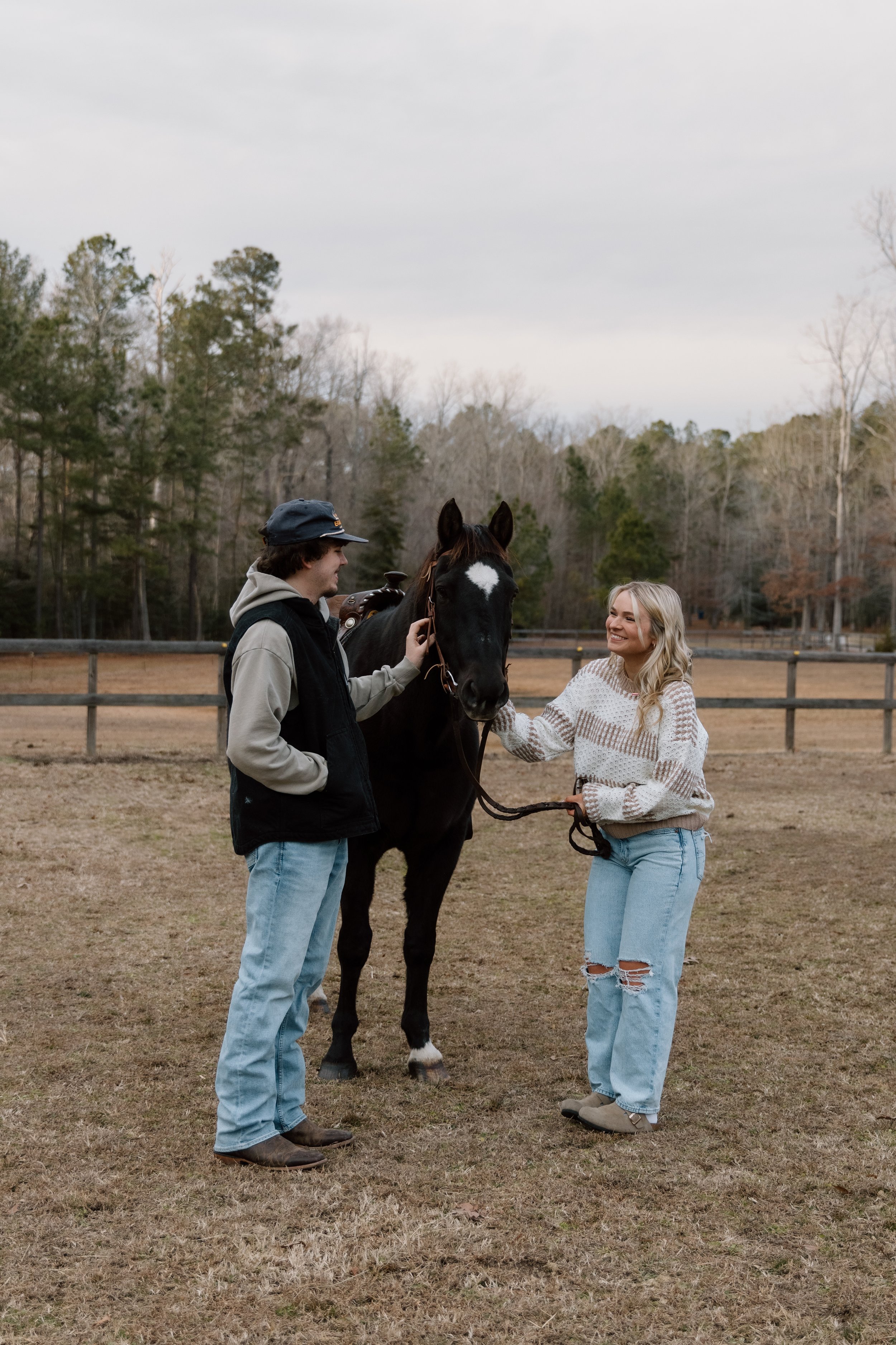 A young man and woman are standing in a field with a black horse, smiling and interacting.