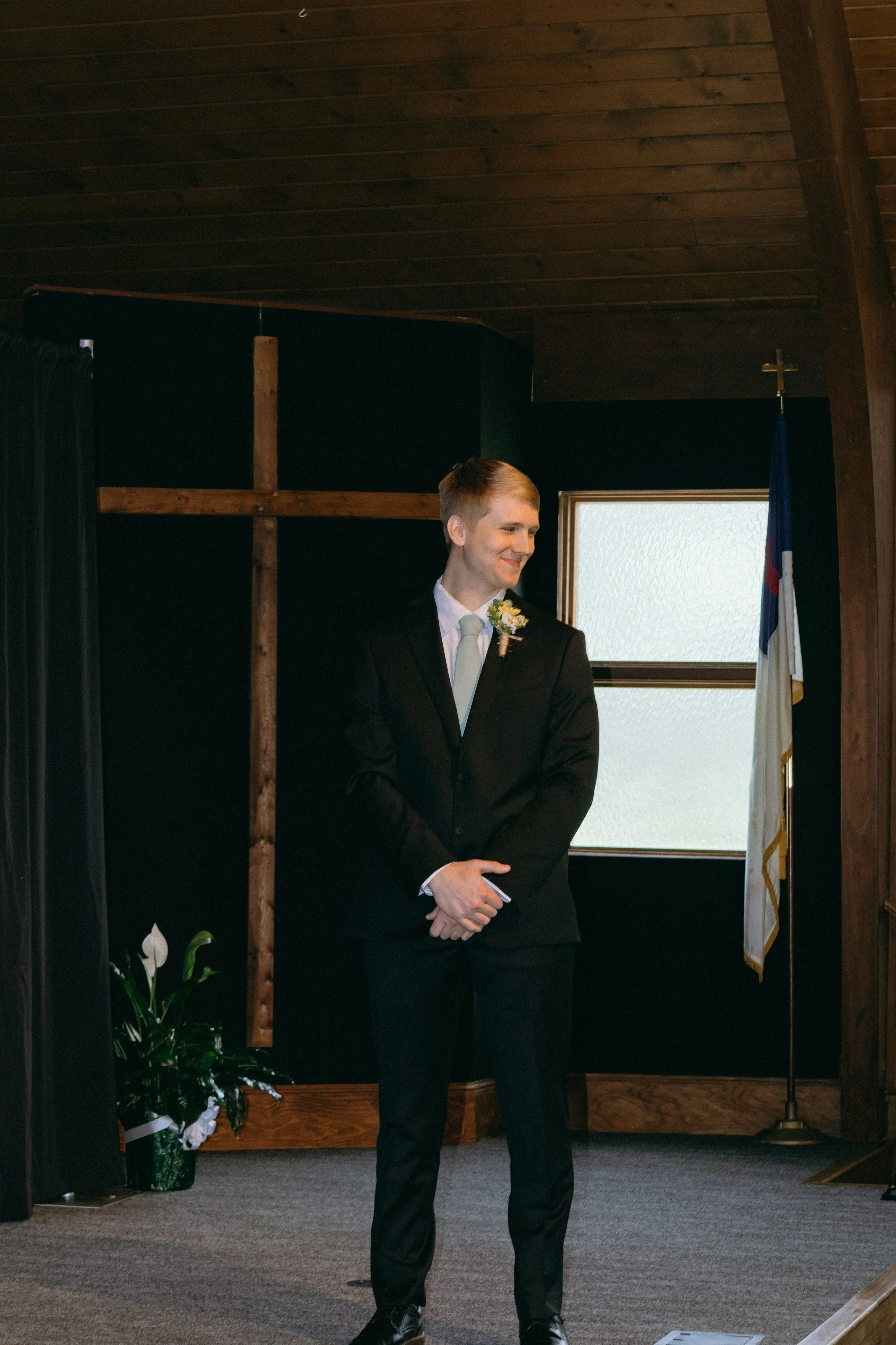 A groom waitng during his wedding ceremony inside a church with a wooden ceiling, cross on the wall, and an American flag.