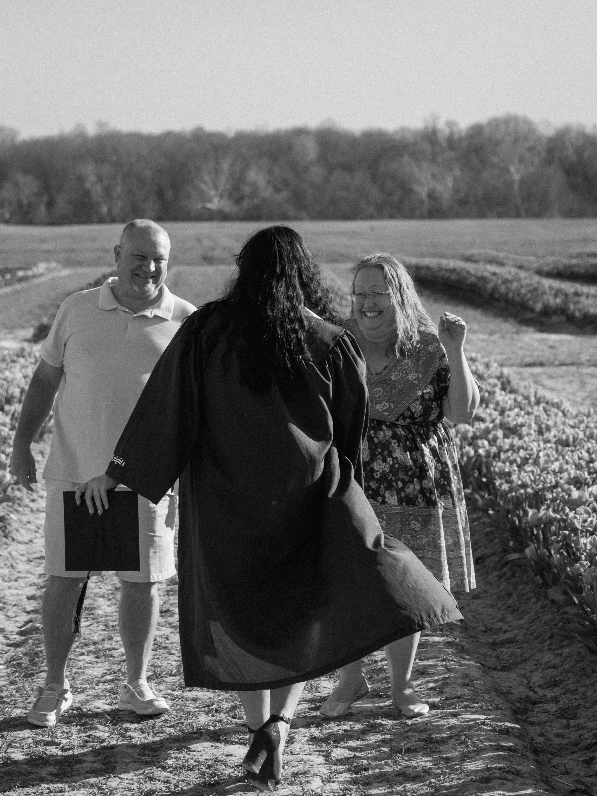 A woman in graduation gown and heels celebrating with a man and woman on a farm field.