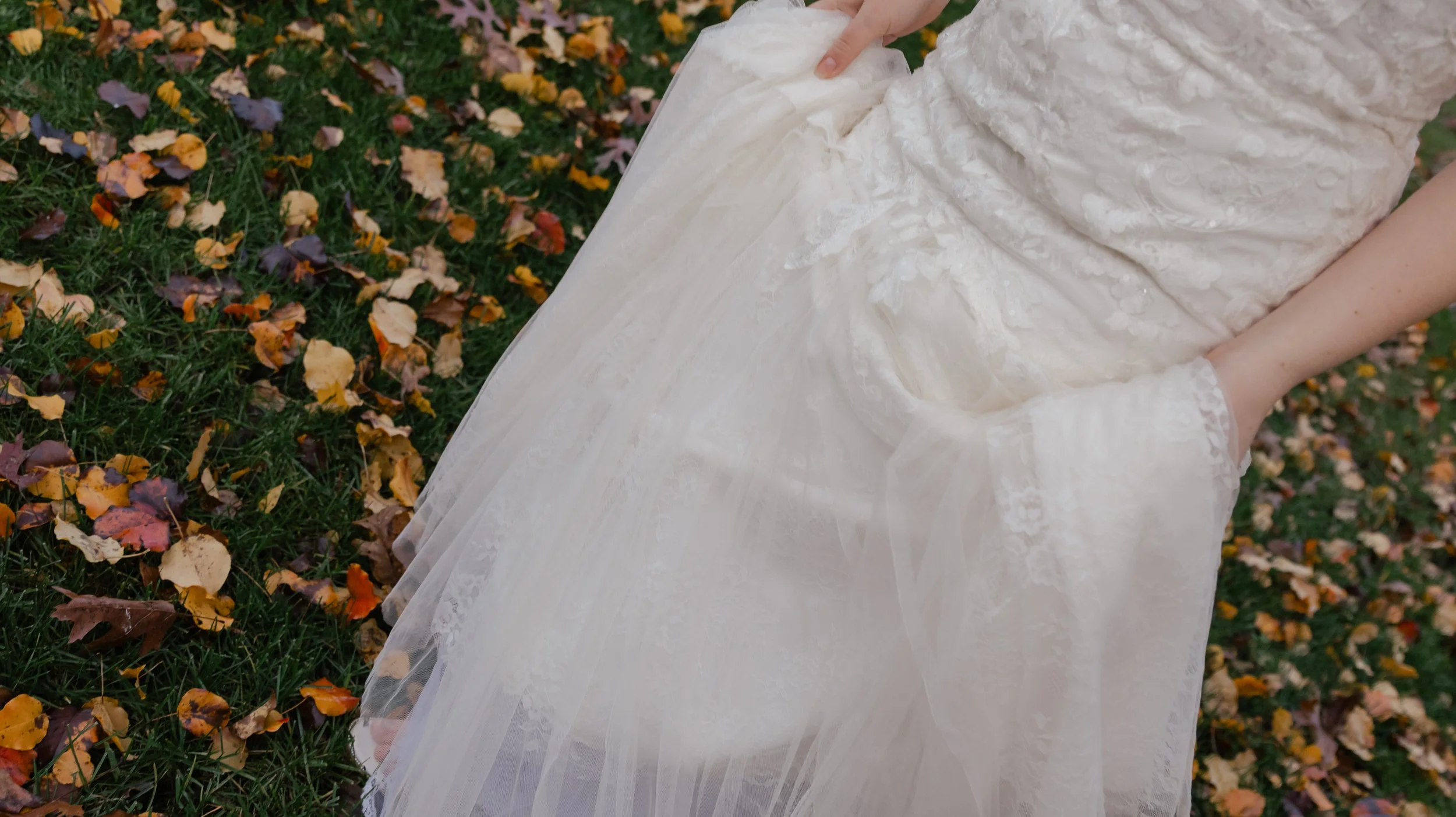 Close-up of a person in a white lace wedding dress with their hand in their pocket, standing on fallen autumn leaves in a grassy area.